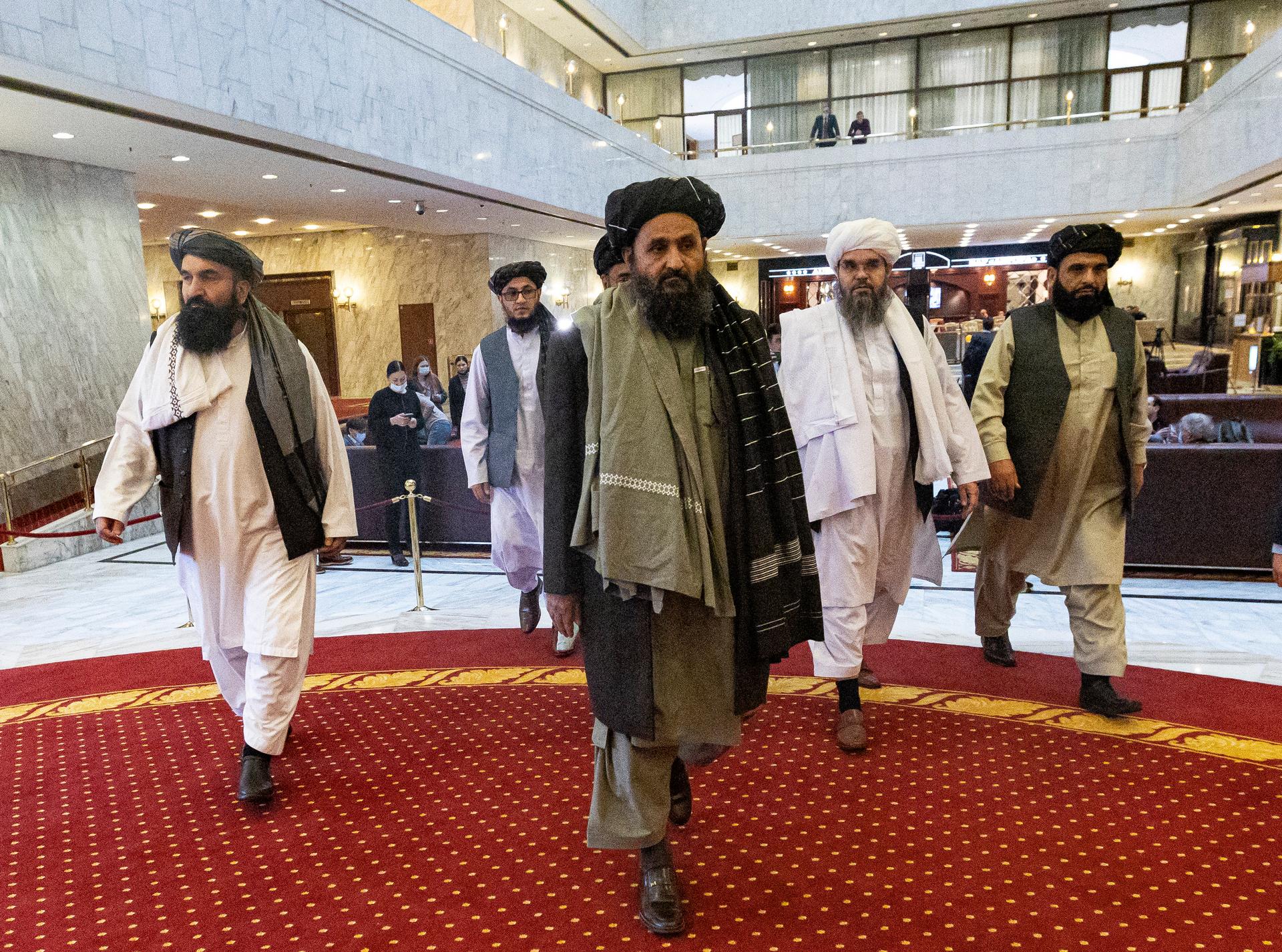 A group of men wearing Afghan traditional clothes walk across a reddish carpet in a lobby.