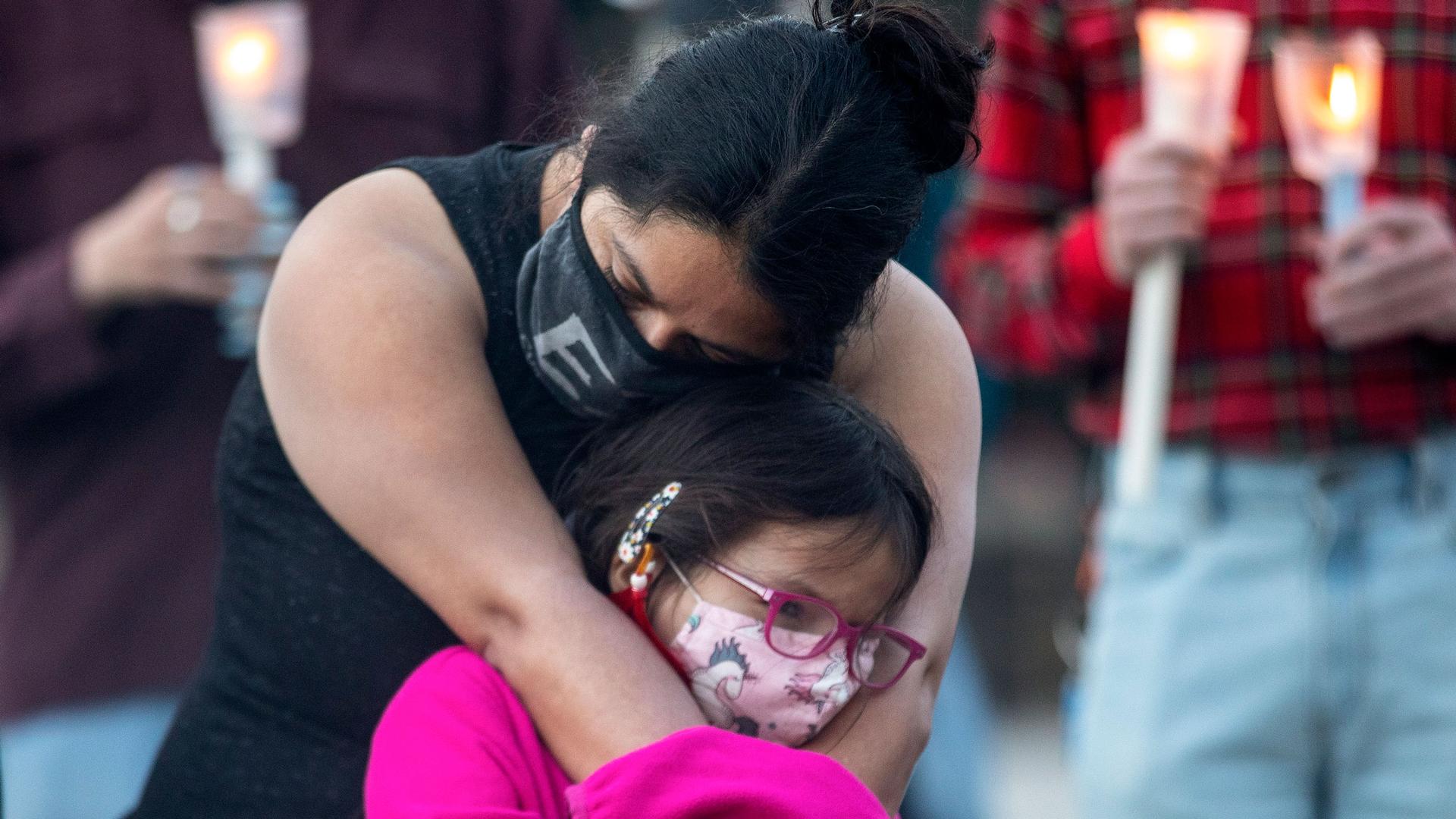 A woman is shown wearing a dark shirt and face mask while hugging a young girl who is wearing a pink shirt.