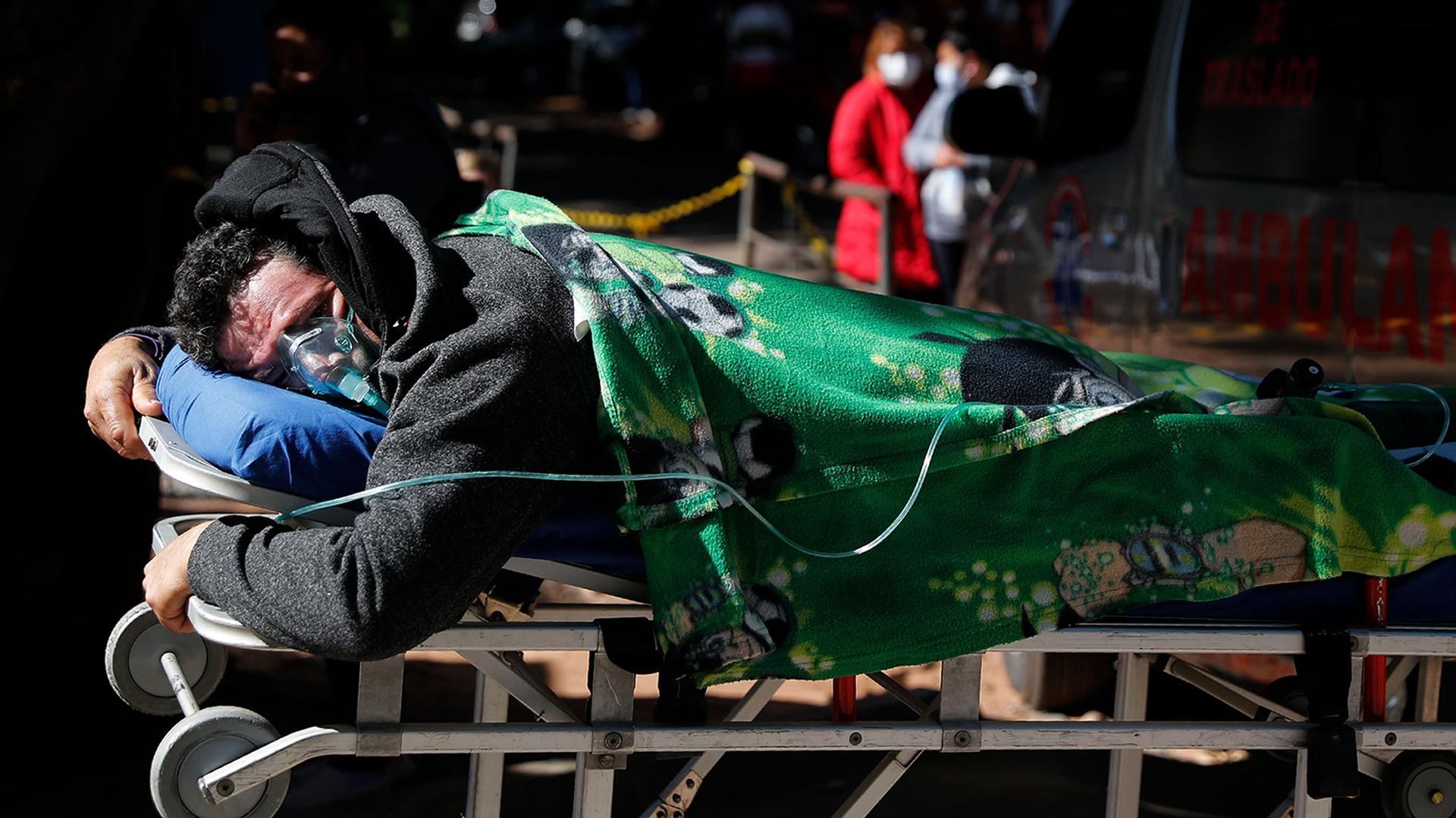 Man with oxygen mask lies face down on a stretcher with a green blanket on top and two people in the distance