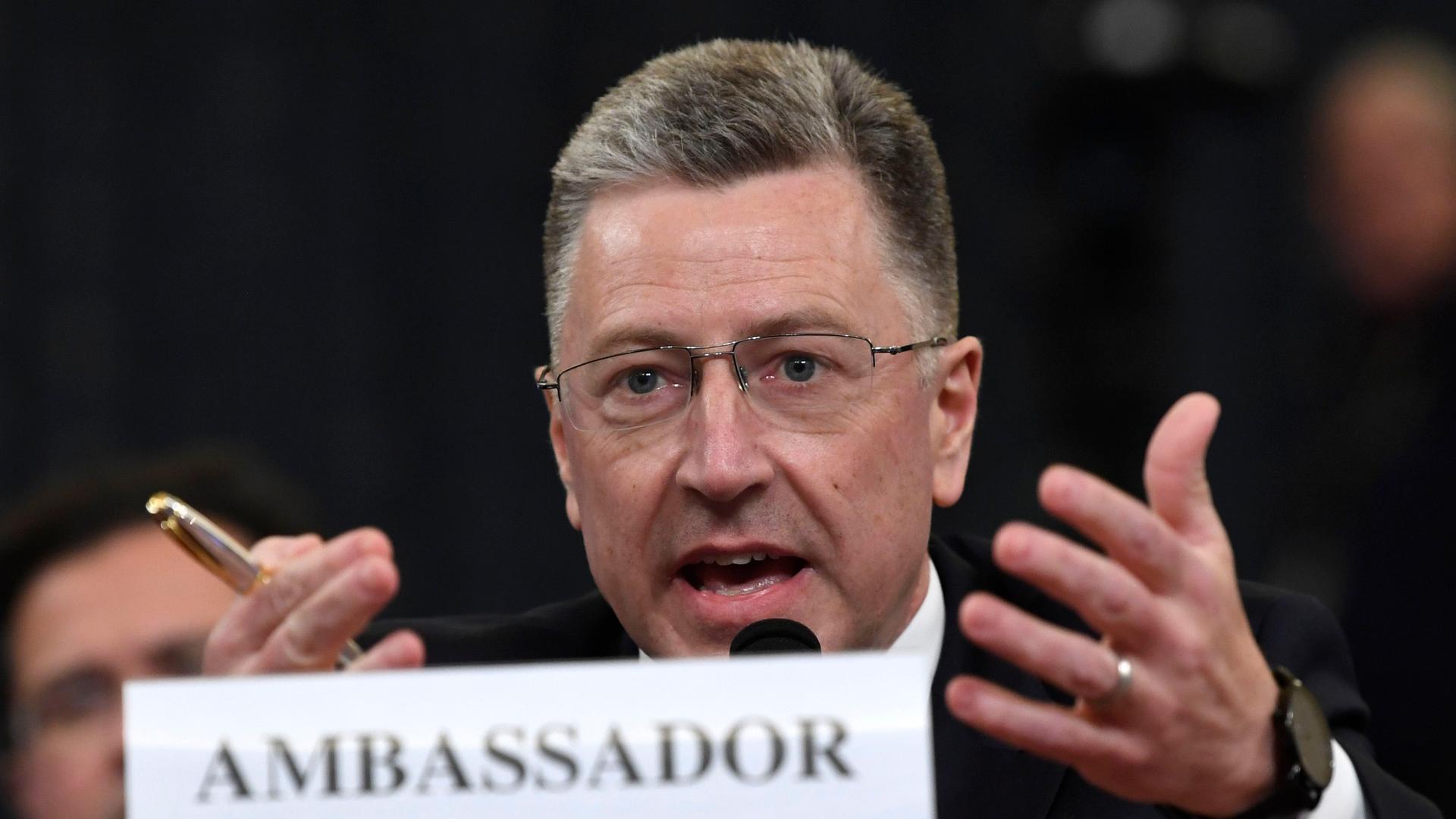A white man wearing glasses with short hair gestures with his hands while speaking with an "ambassador" sign.