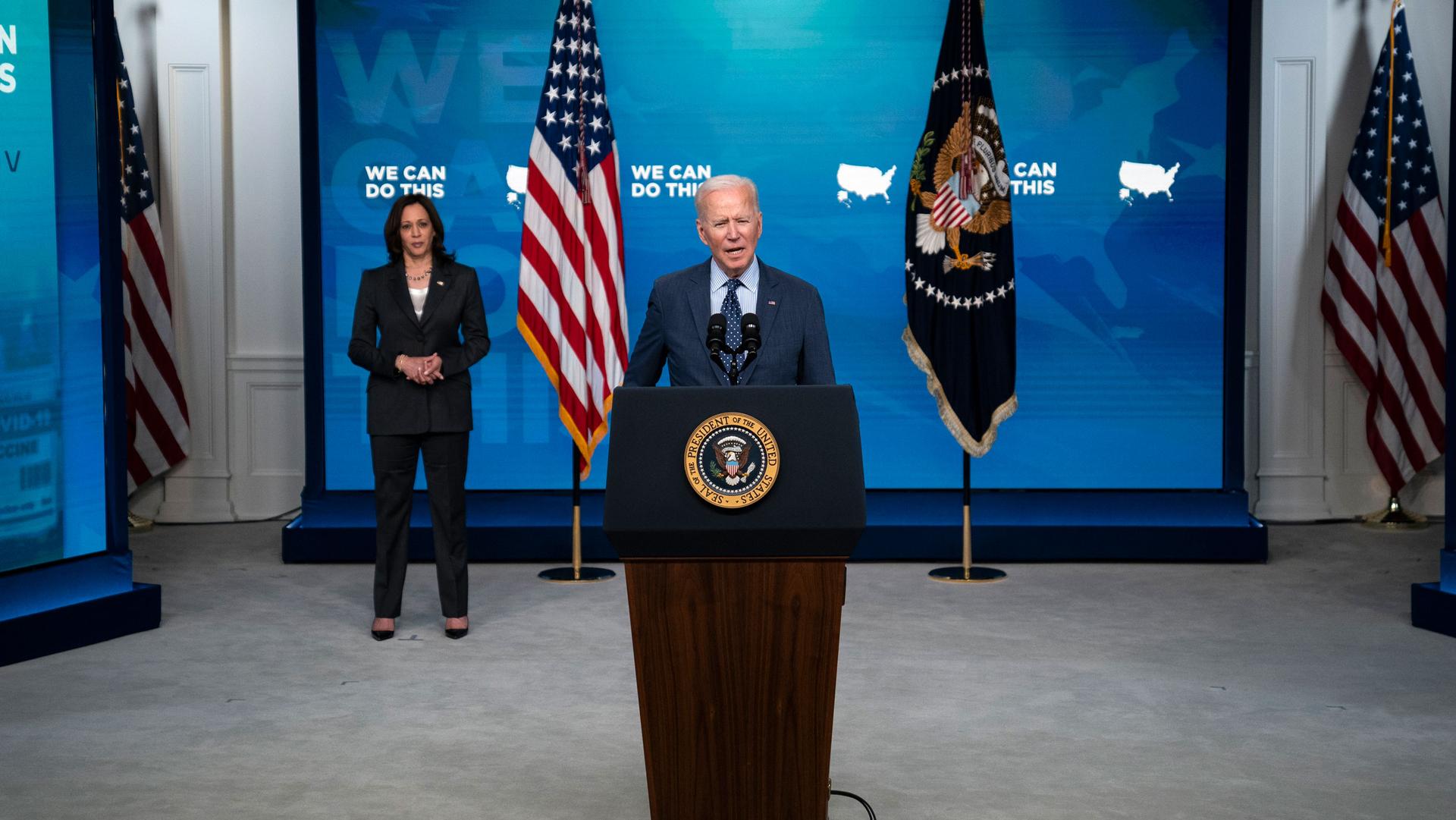 Vice President Kamala Harris is shown standing several feet behind President Joe Biden who is standing at a wooden podium.