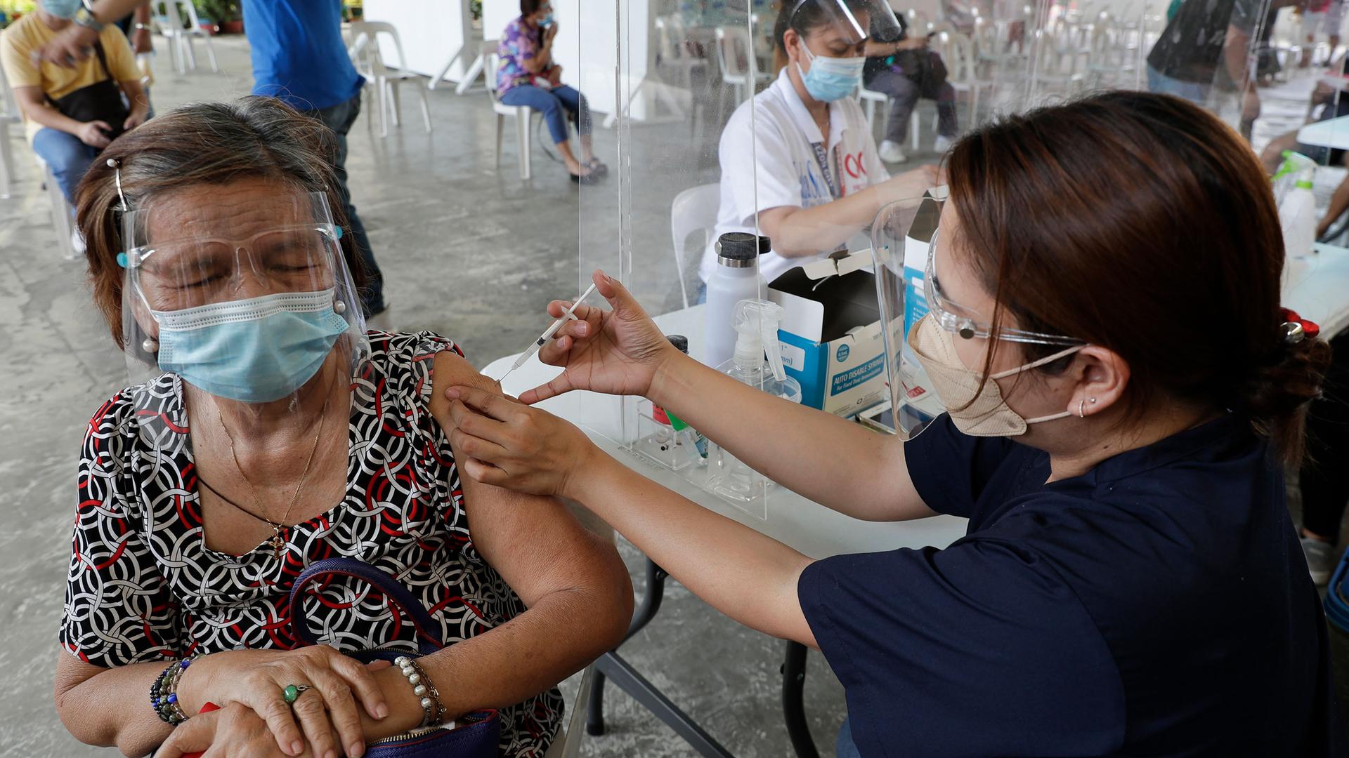 A woman is shown with a clear face shield and face mask while receiving a vaccination shot by a health worker.