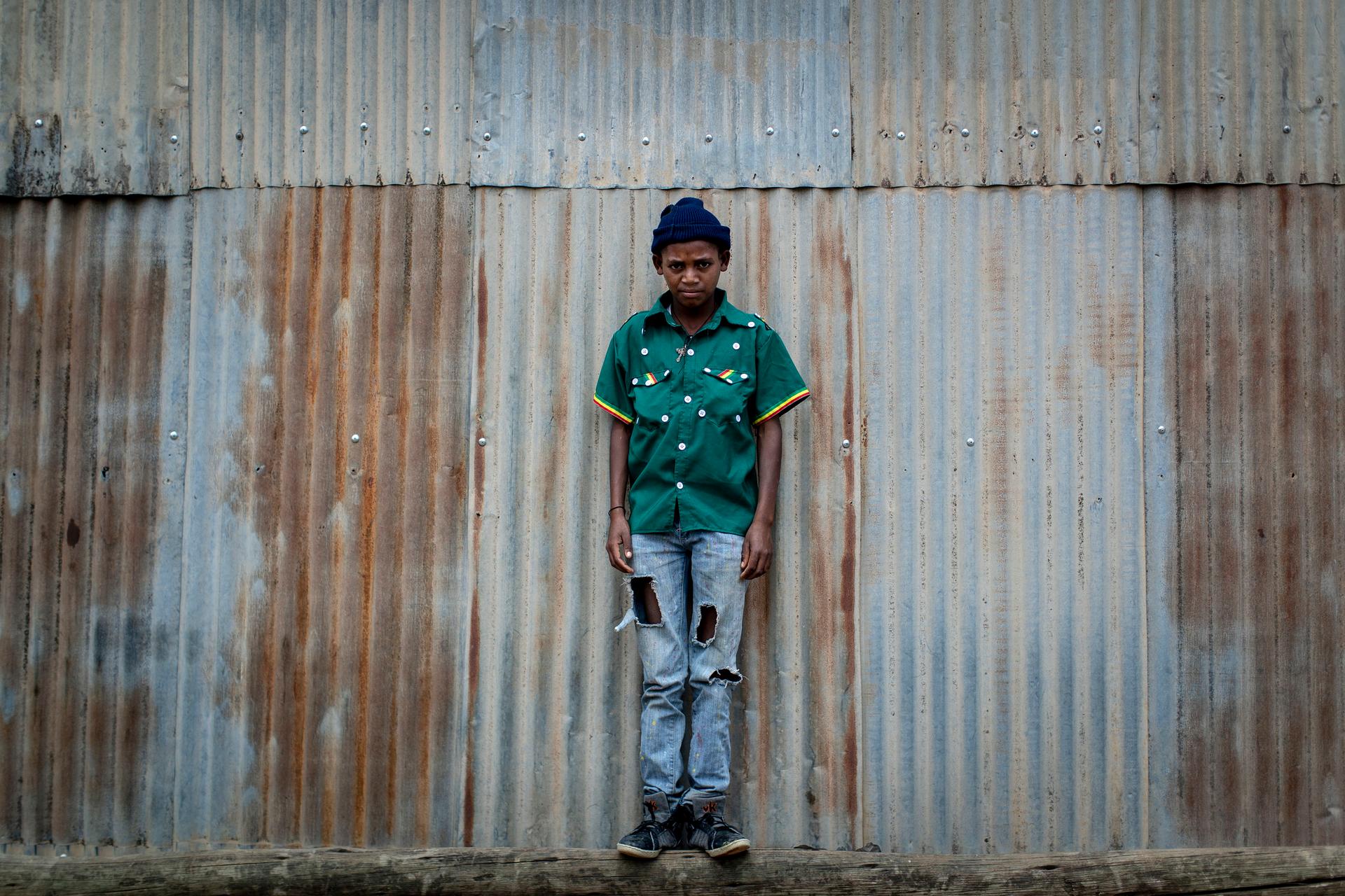 boy stands against corrugated steel wearing a green cultural shirt with buttons and jeans.