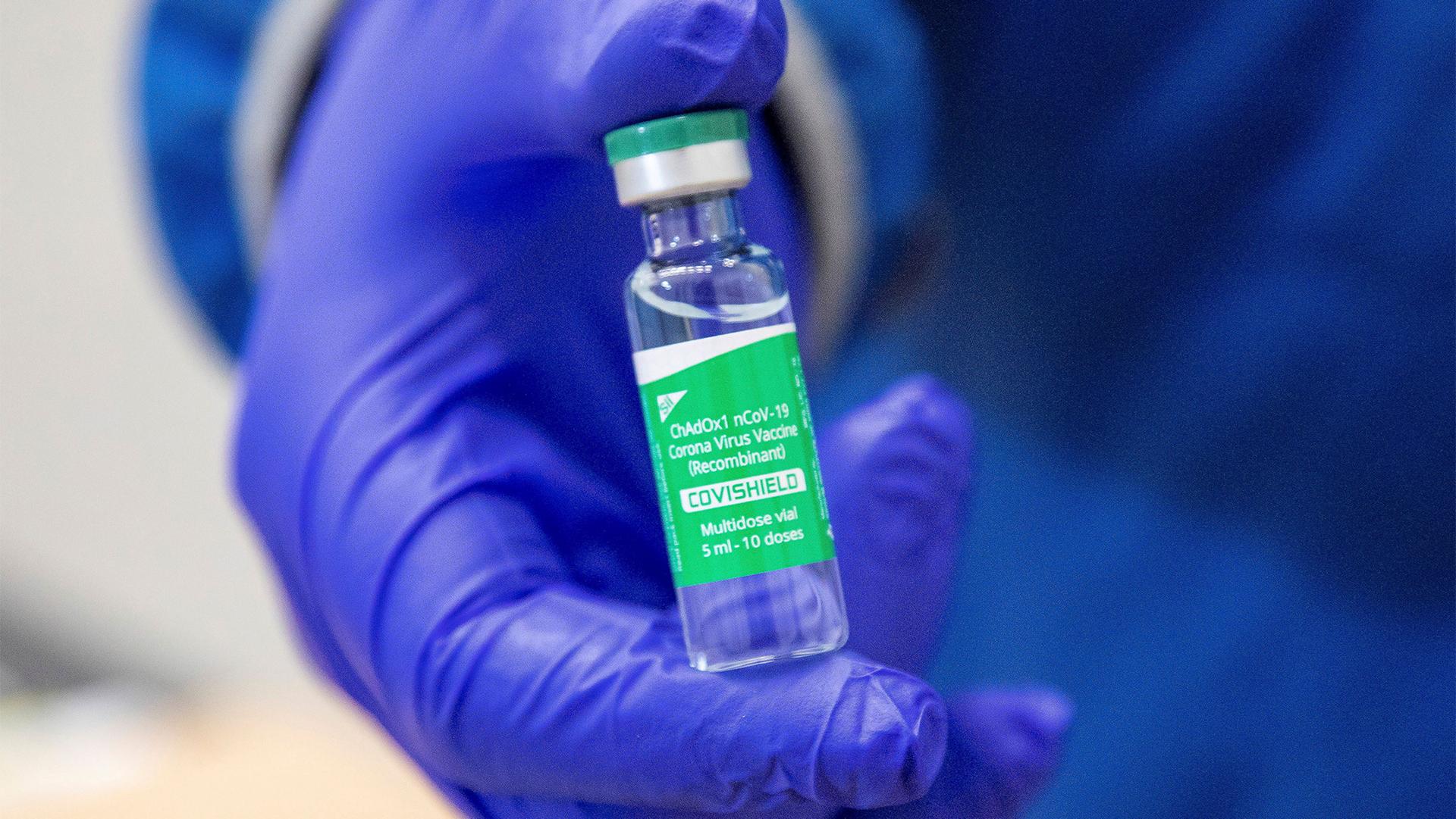 A person wearing blue gloves presents a vial of an AstraZeneca COVID-19 vaccine dose