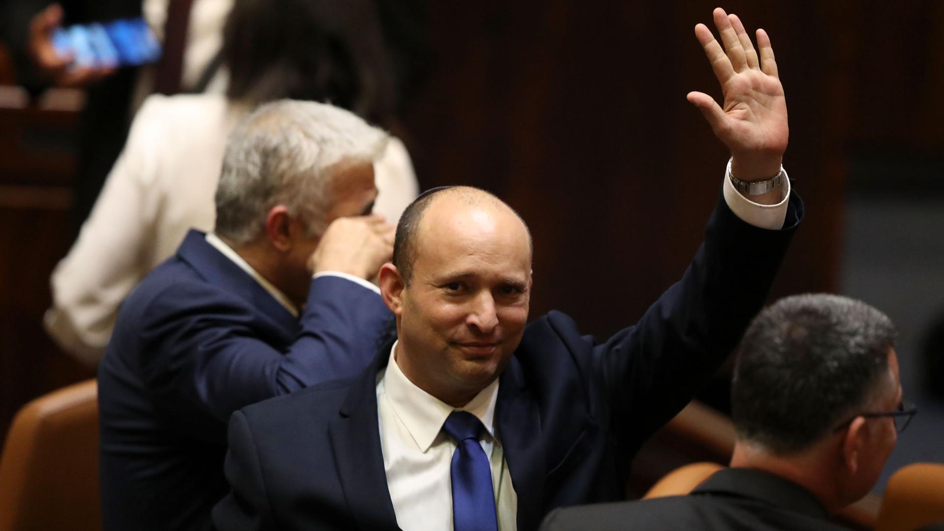 Naftali Bennett is shown wearing a dark suit and raising his right hand in the air.