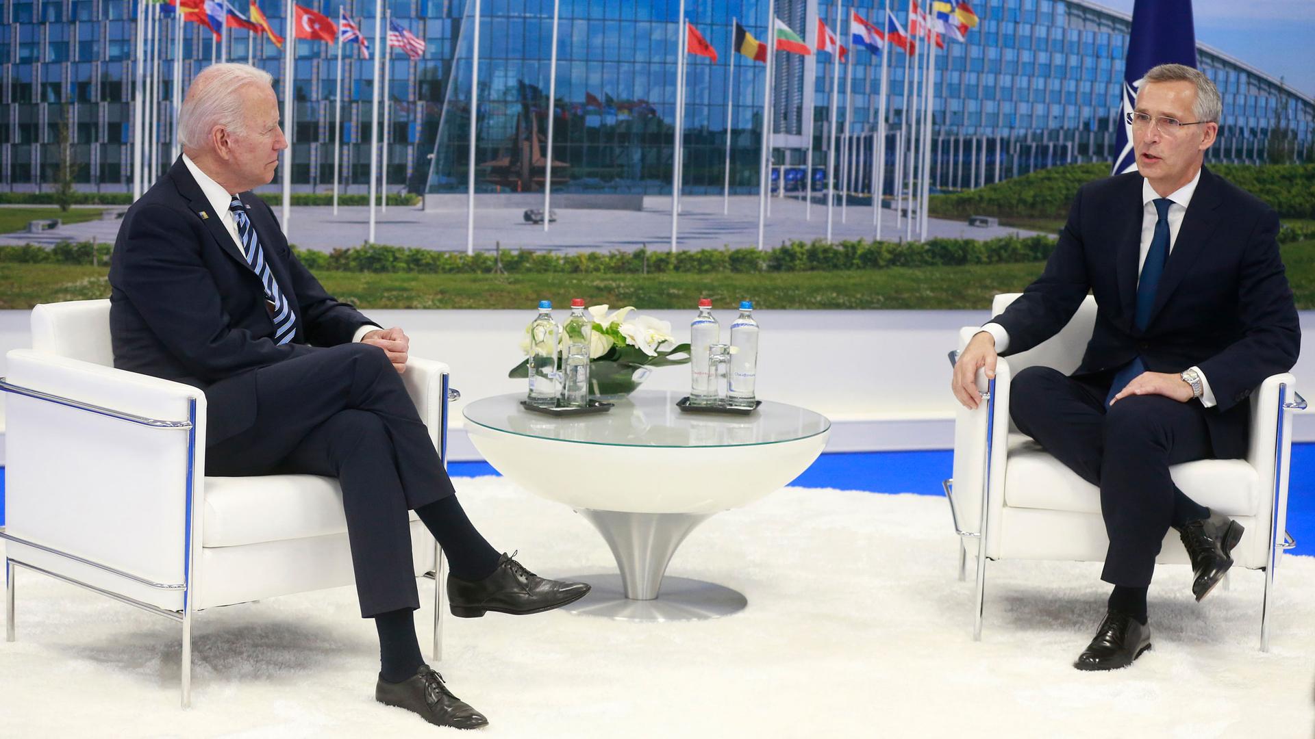 NATO Secretary General Jens Stoltenberg and US President Joe Biden are shown both wearing suits and sitting in white armchairs.