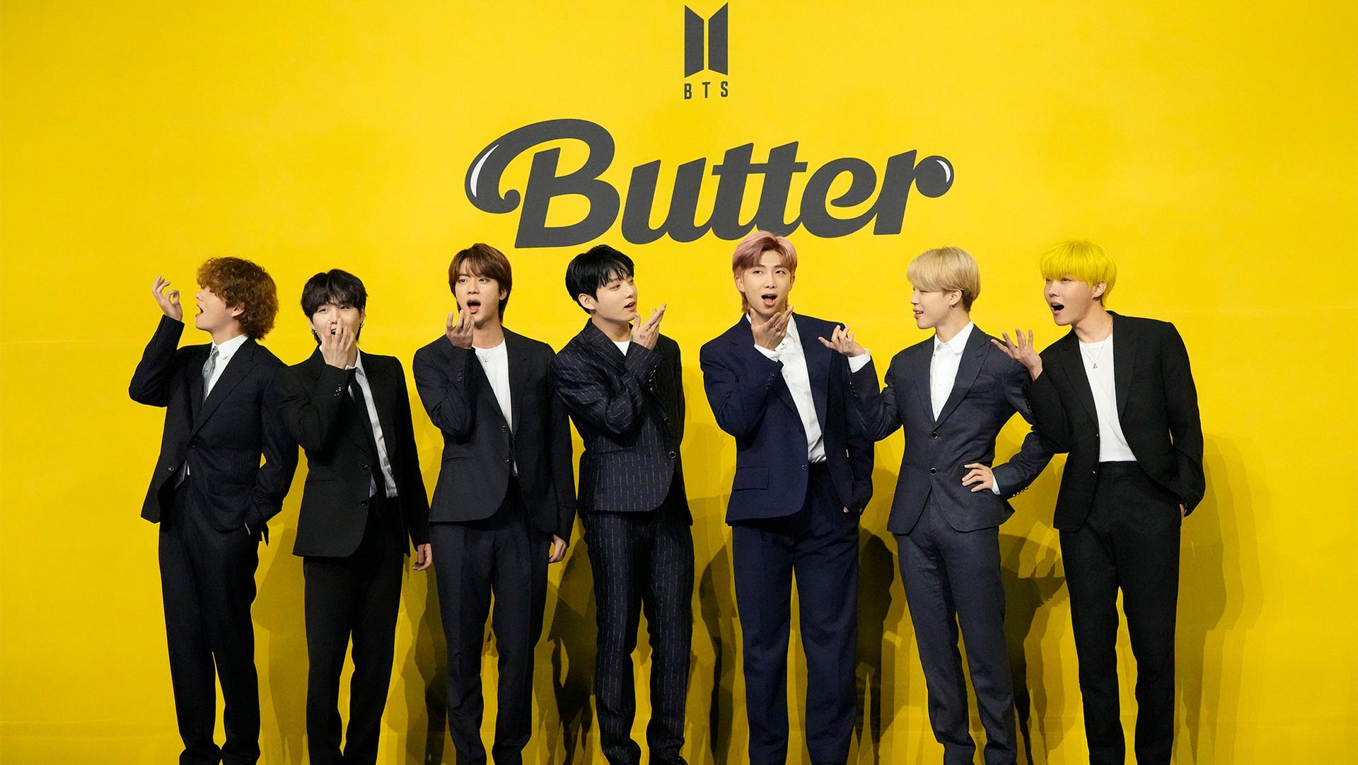 Seven members of K-pop band BTS stand wearing black suits in front of a yellow background that says "BTS Butter"