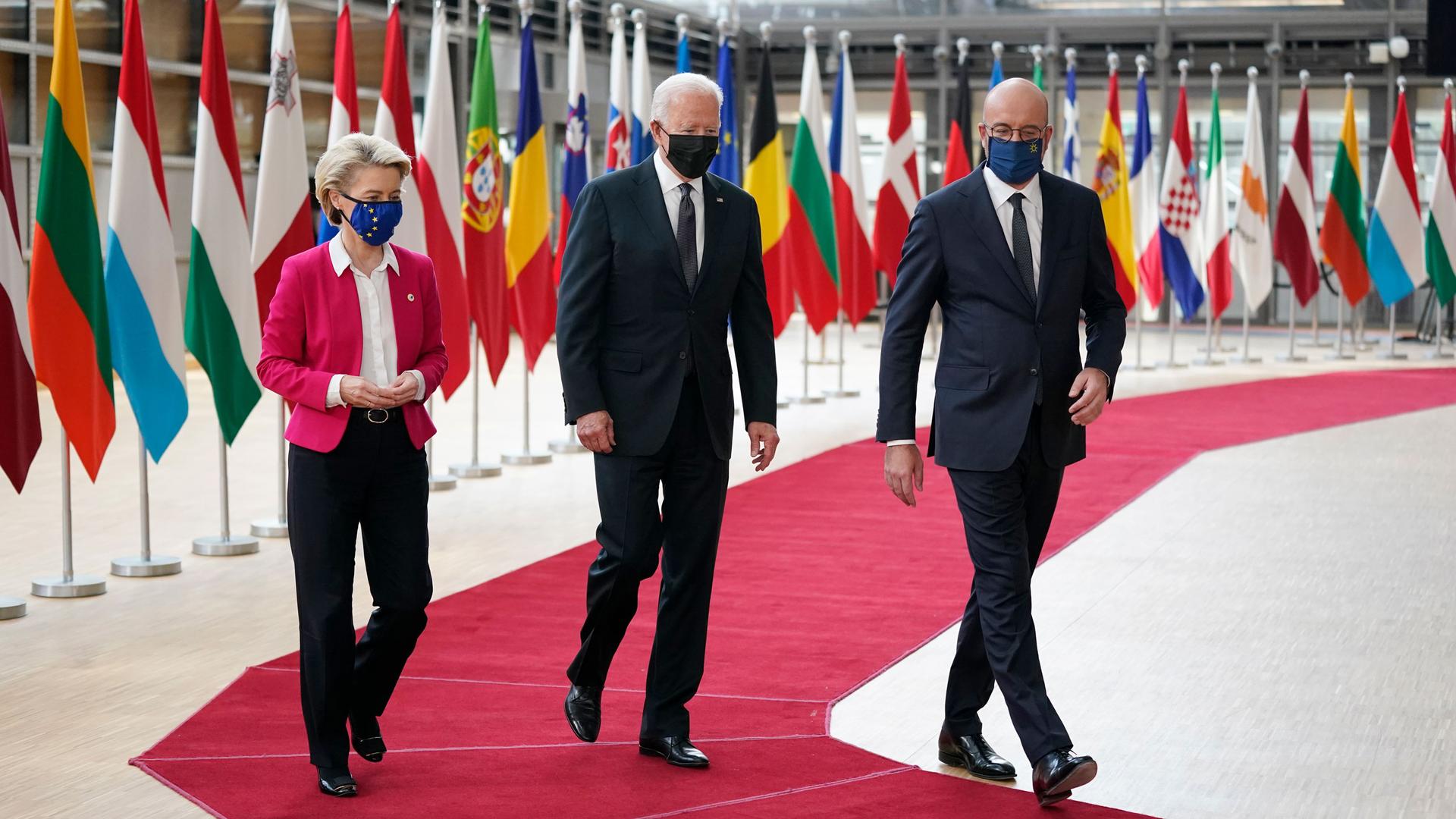 Joe Biden, Charles Michel and Ursula von der Leyen are shown walking side-by-side on a red carpet with several national flags in the background.