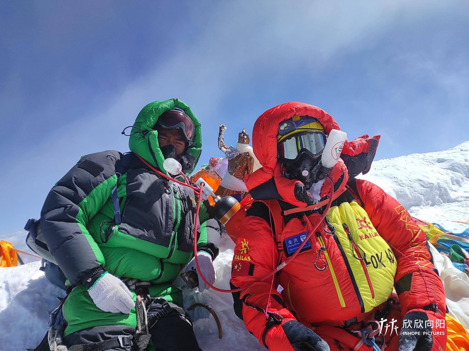 Two men wearing green and red coats stand amid a snowy Mount everest