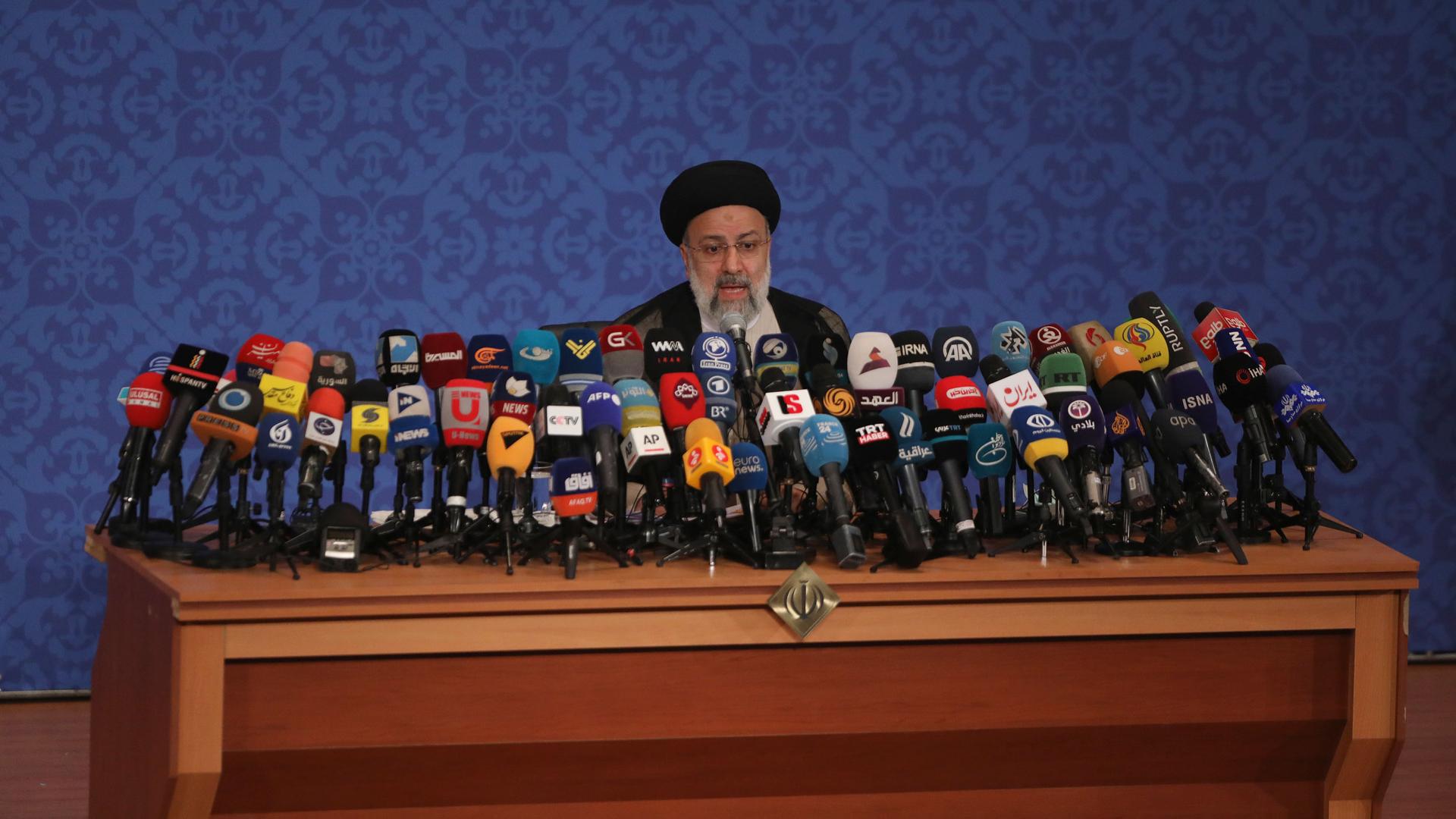 Iran's new President-elect Ebrahim Raisi is shown standing at a podium with several dozen microphones.