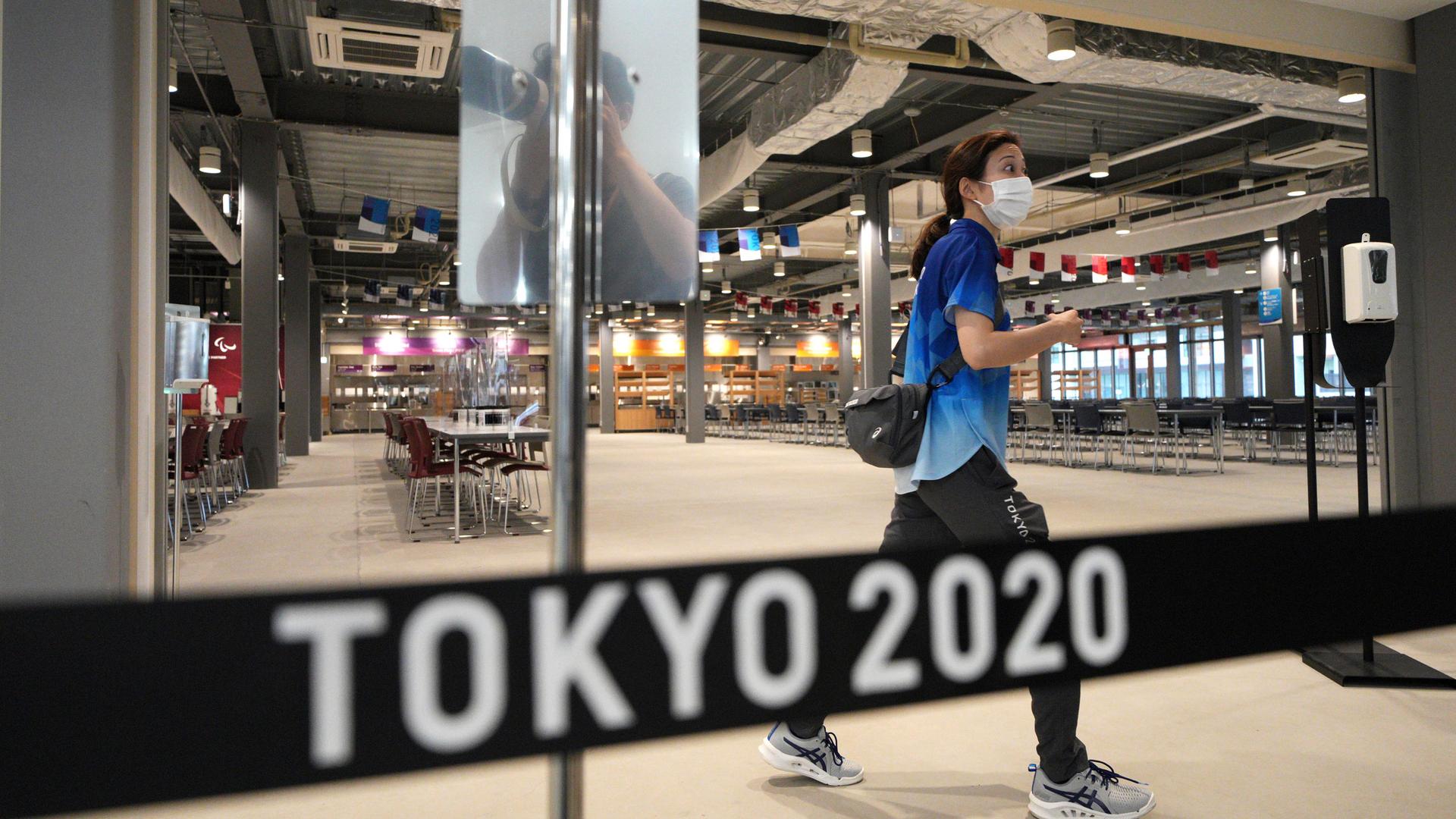 A woman is shown walking and wearing a face mask and blue t-shirt in a nearly empty dining hall with a sign in the nearground that says "Tokyo 2020."