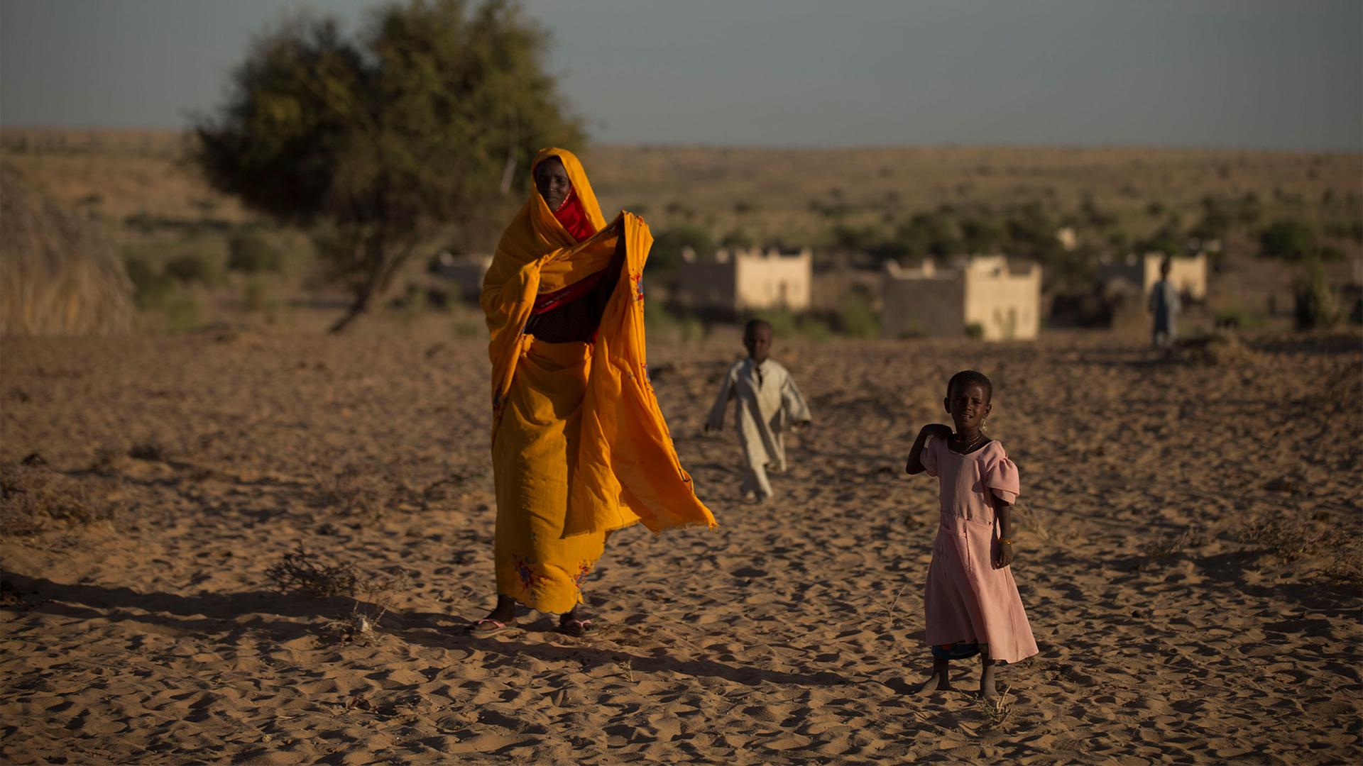 Woman in orange outfit walks in dry climate with girl in pink dress and boy in beige outfit