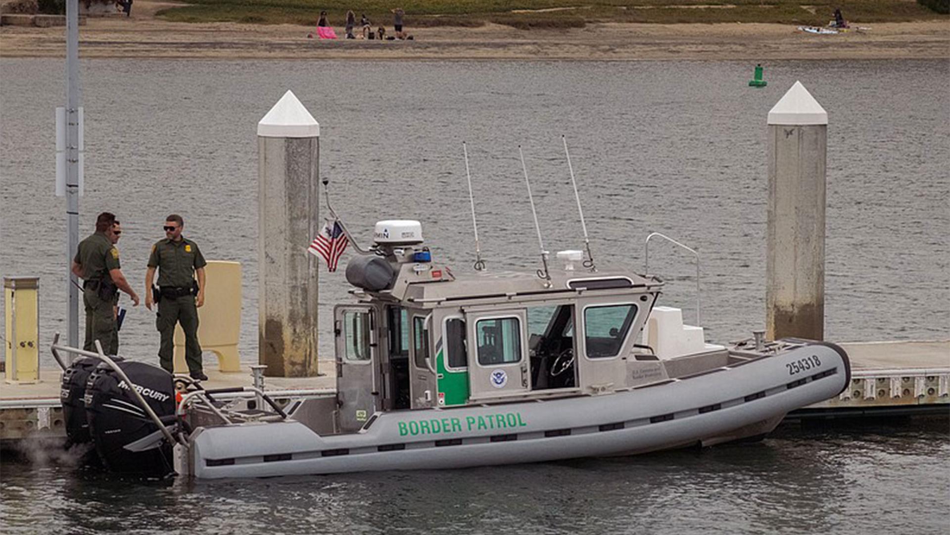 Three men in green suits stand on a small boat that says "Border Patrol" with an American flag in the middle