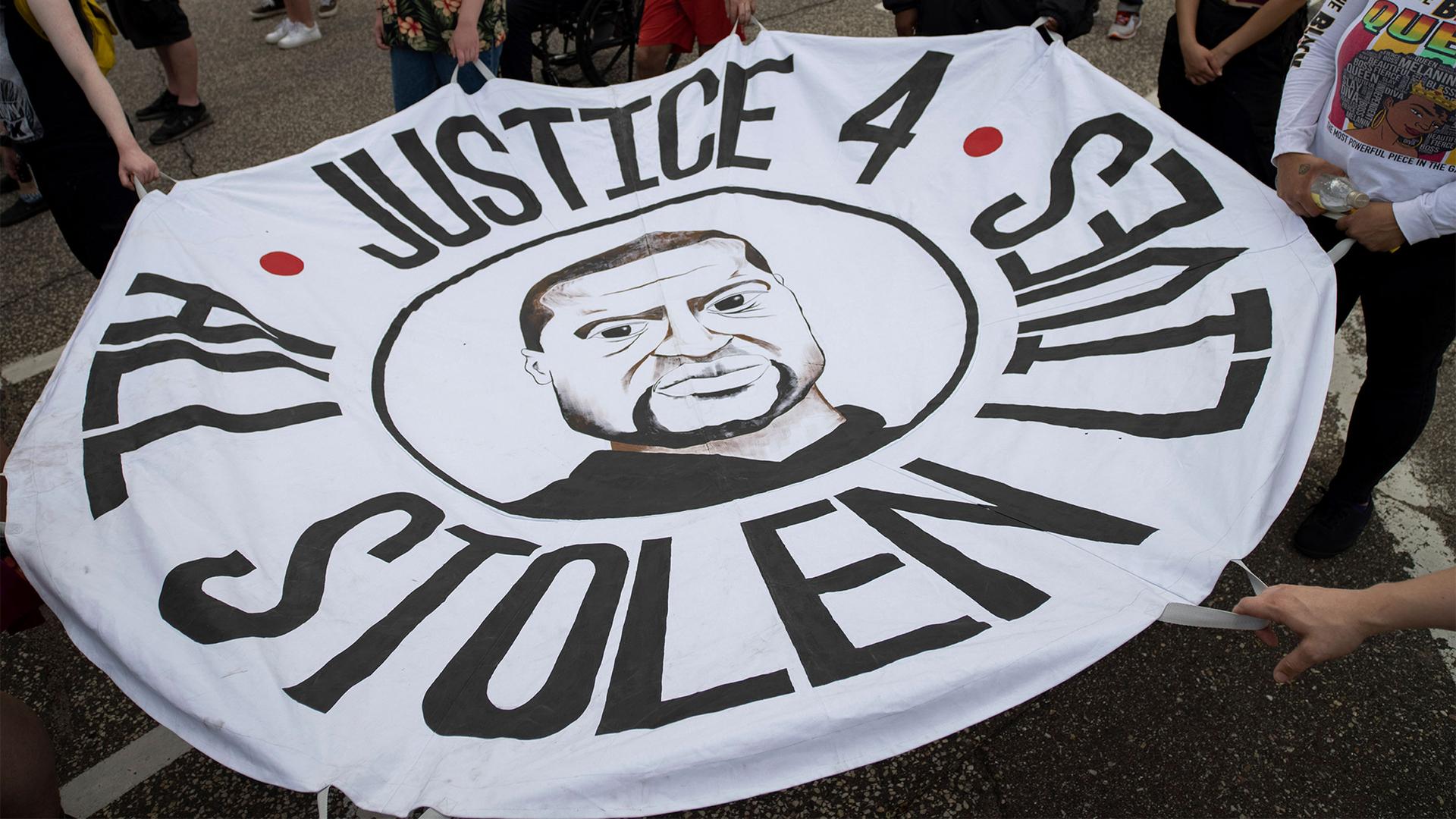 Hands hold a round white cloth circle with George Floyd's face in the middle that reads, "Justice 4 all stolen lives"