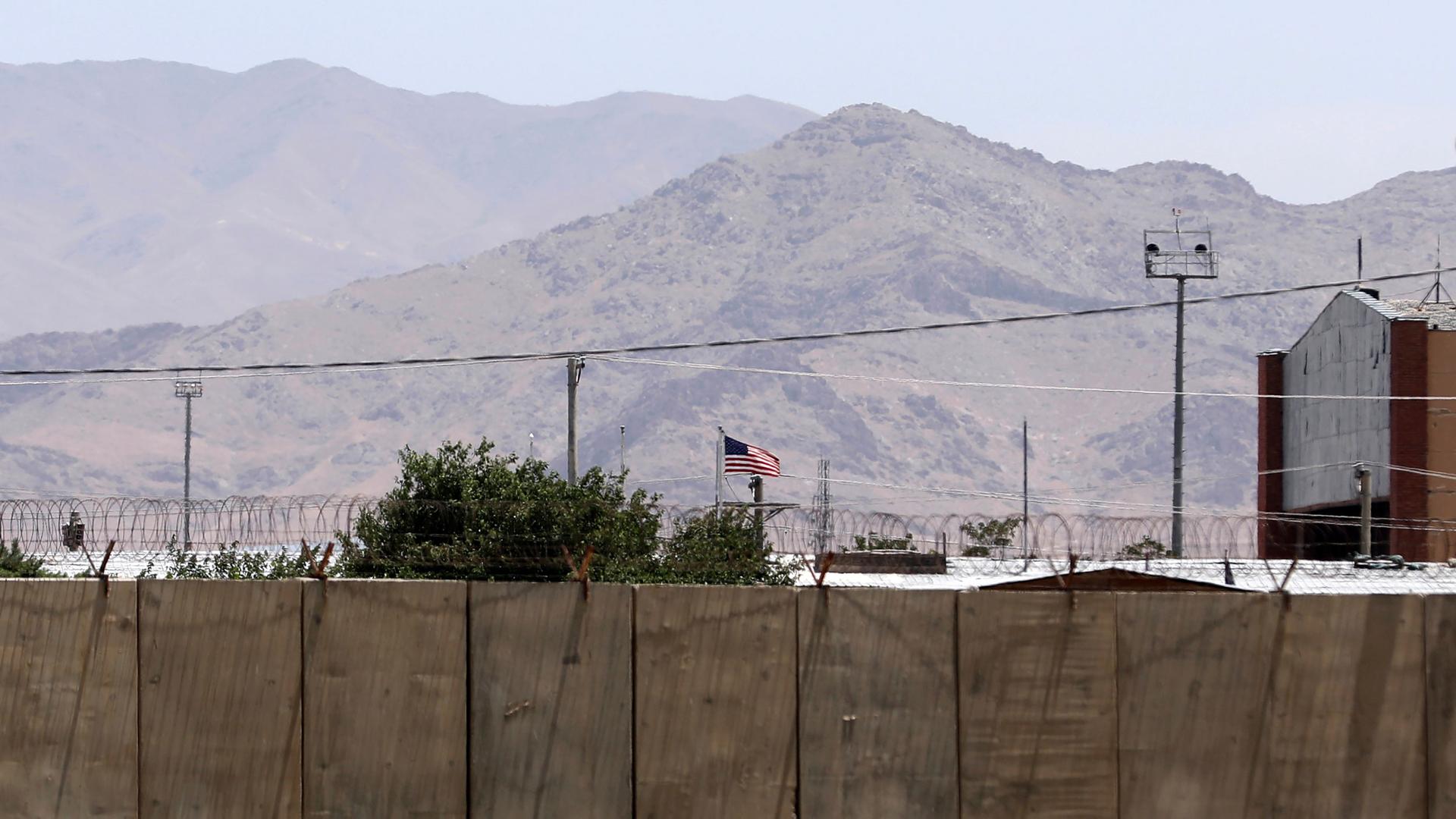 A large security wall is shown in the nearground with the US flag flying and a mountain range in the distance.