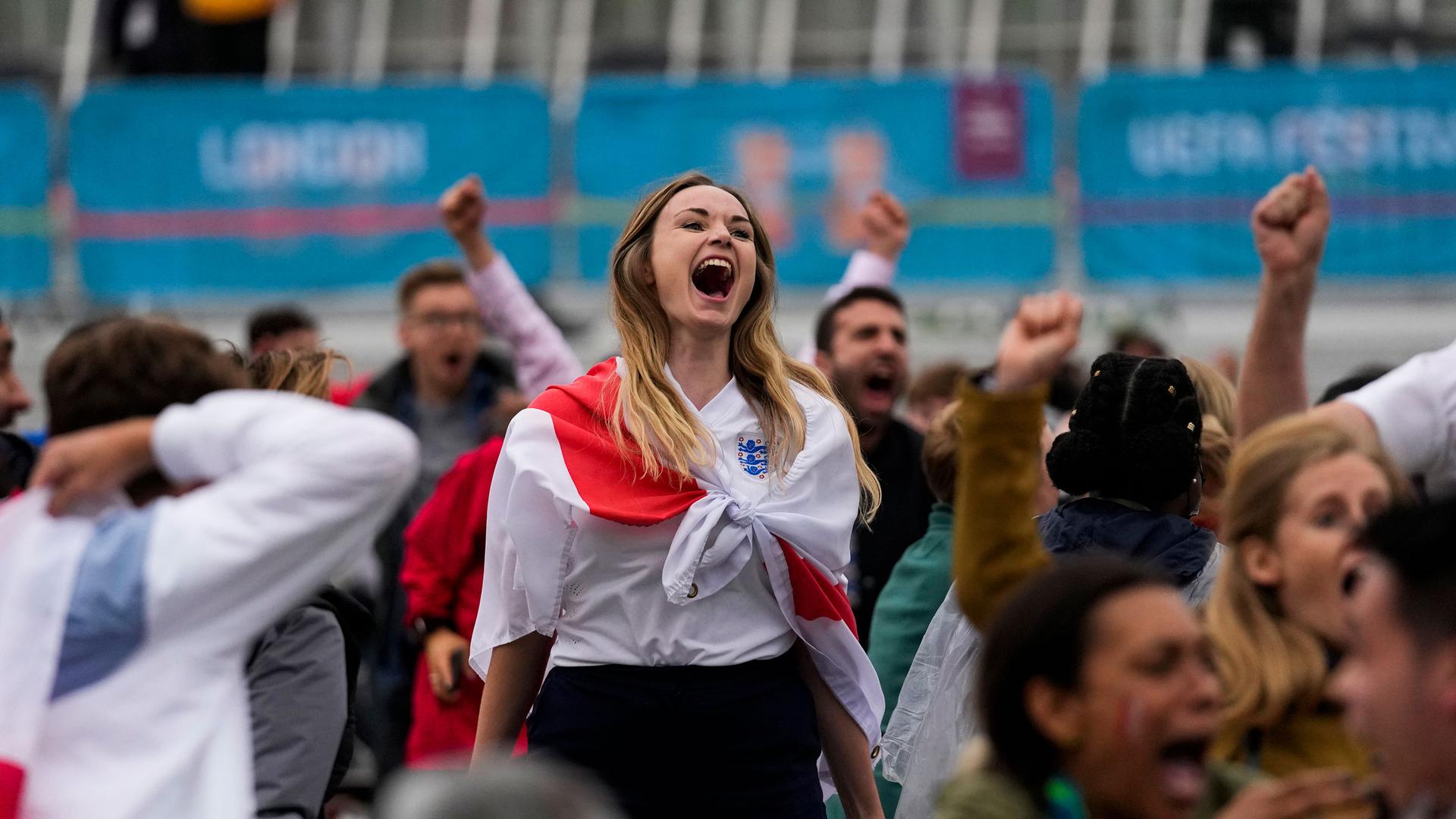 A large crowd of people are shown with a woman in the center of the photograph cheering with her mouth open and an England flag draped on her shoulders.