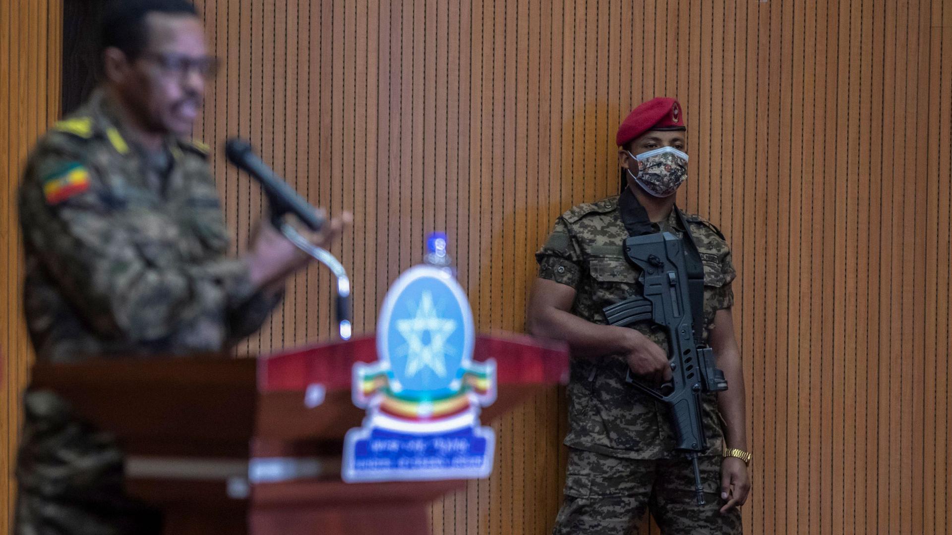 Lieutenant-General Bacha Debele is show standing at a podium with his hand raised in soft focus with a guard in military fatigues is shown in focus and holding a large rifle. 
