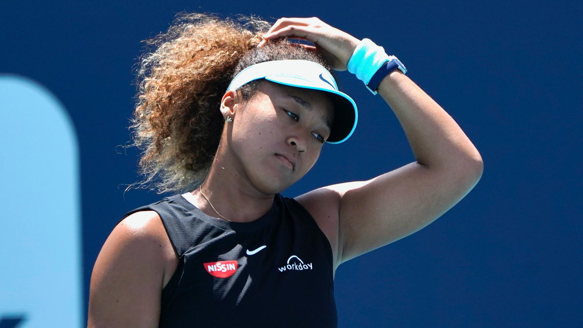 Naomi Osaka, in a tennis hat and ponytail, puts her hand on her head