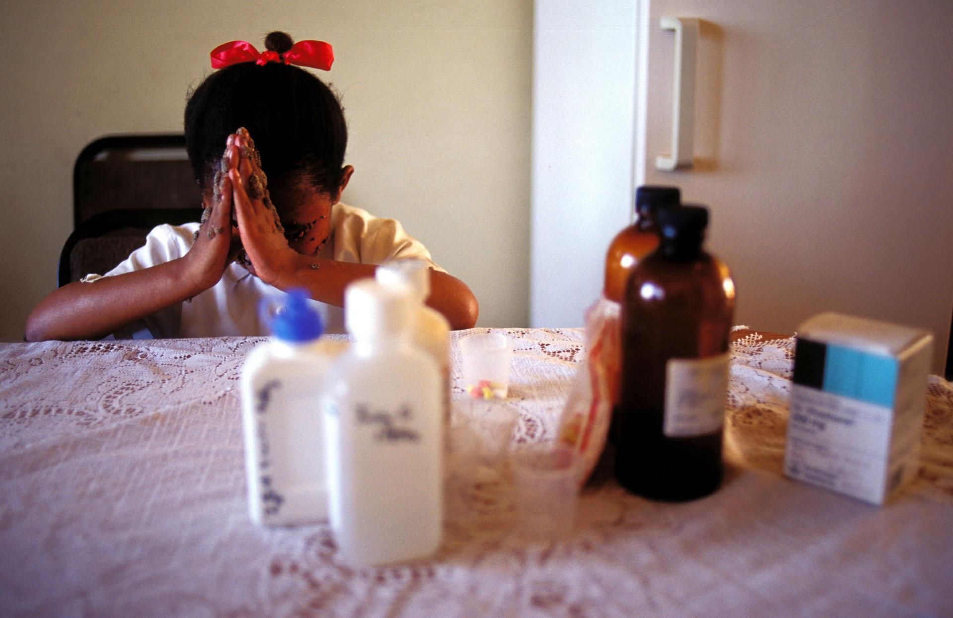 A young girl folds her hands in prayer in front of her. Out of focus, in front of her, is a group of medication bottles.