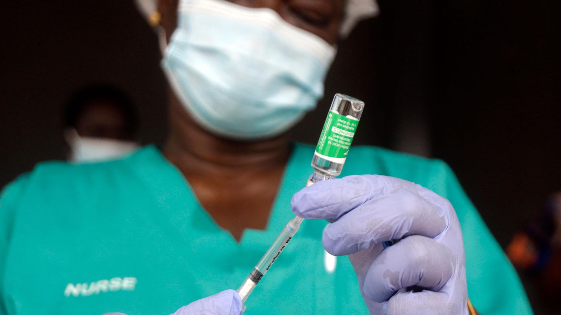 A close up photograph of a syringe is shown with a nurse in soft focus in the background.