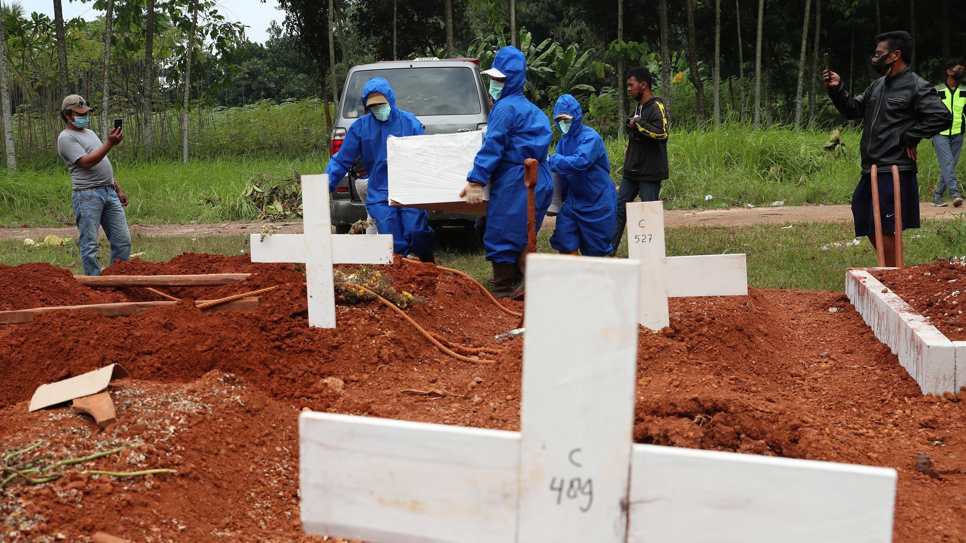 Several workers are shown wearing blue protective suits and carrying a white coffin while walking in at a grave site with several white crosses.
