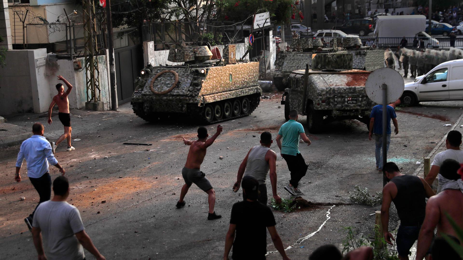 Several people are shown in a street throwing items at a tank and other armored vehicles.