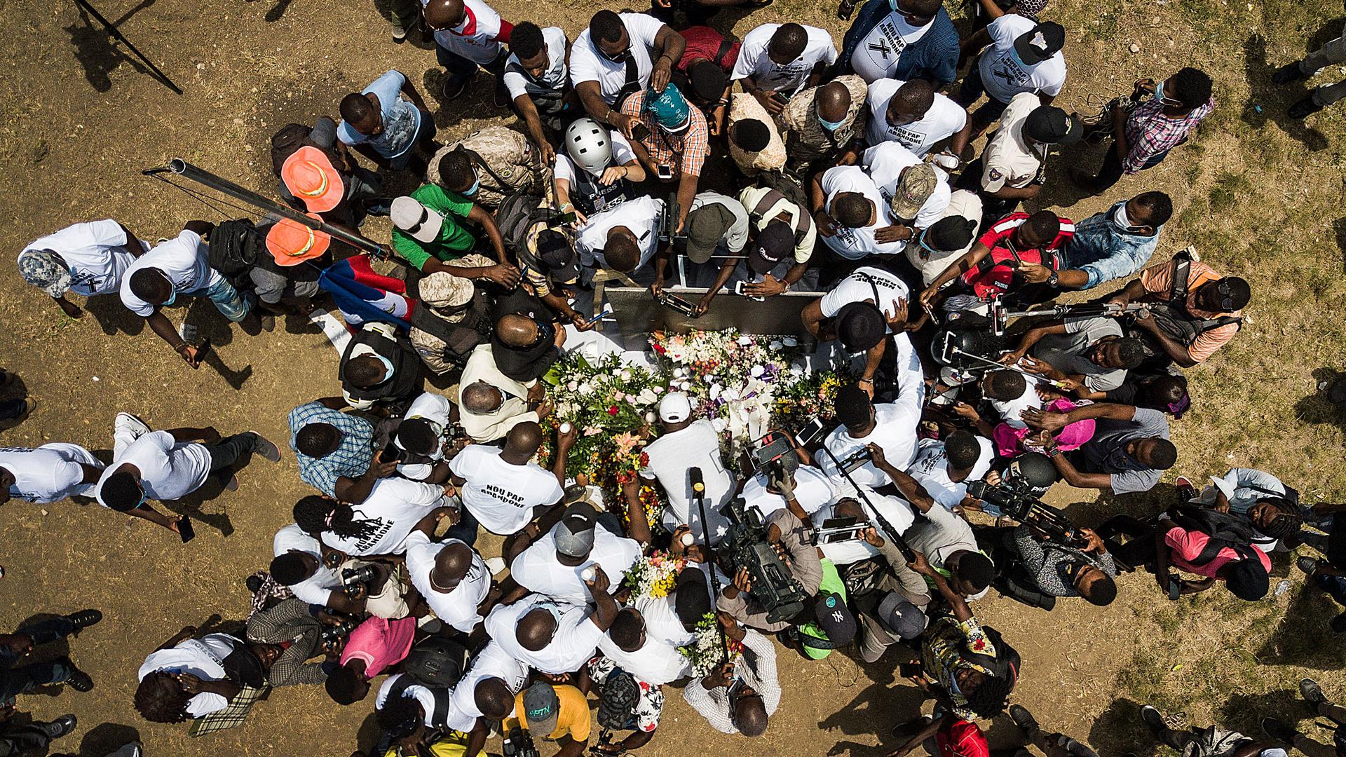 A large group of people are shown from above circling above a memorial of flowers.