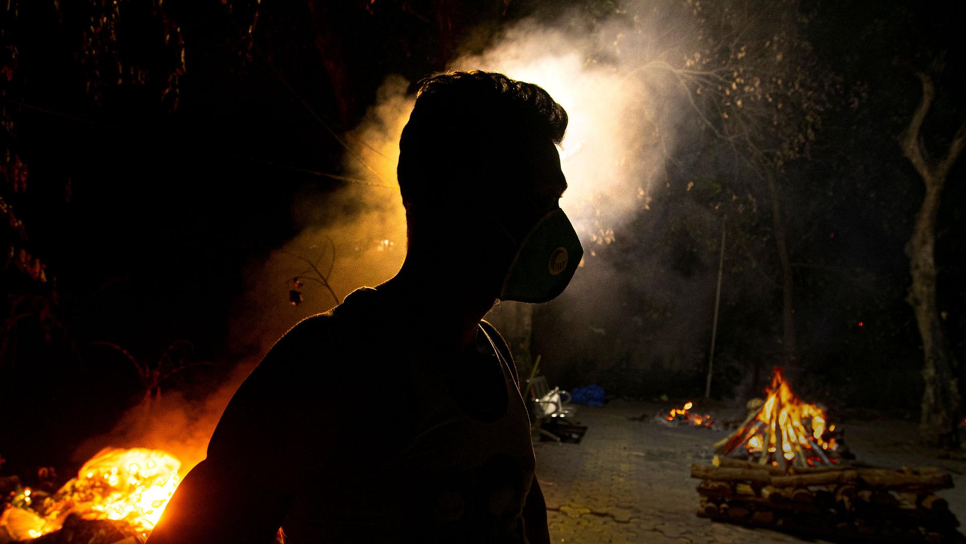 A man is shown in shadow wearing a face mask with several large glowing orange funeral pyres in the distance.