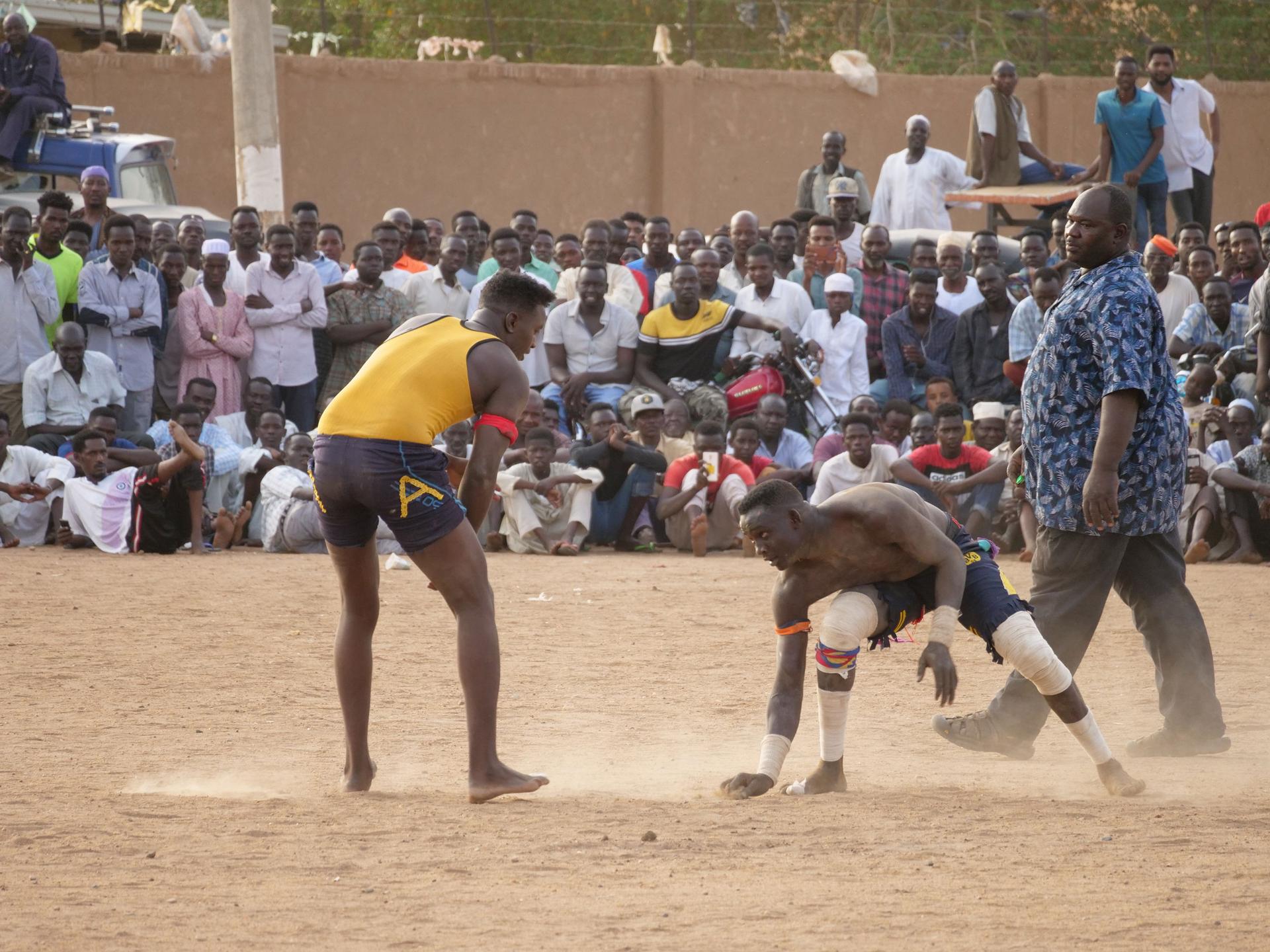 To men are shown wearing shorts and t-shirts while wrestling in a sandy courtyard.