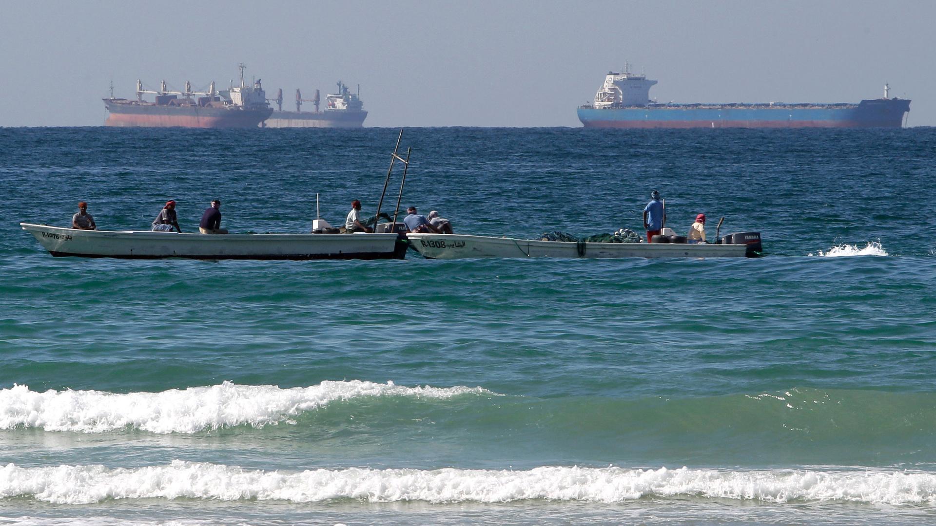 Two small fishing boats are shown in the near ground with large oil tankers off in the distance.