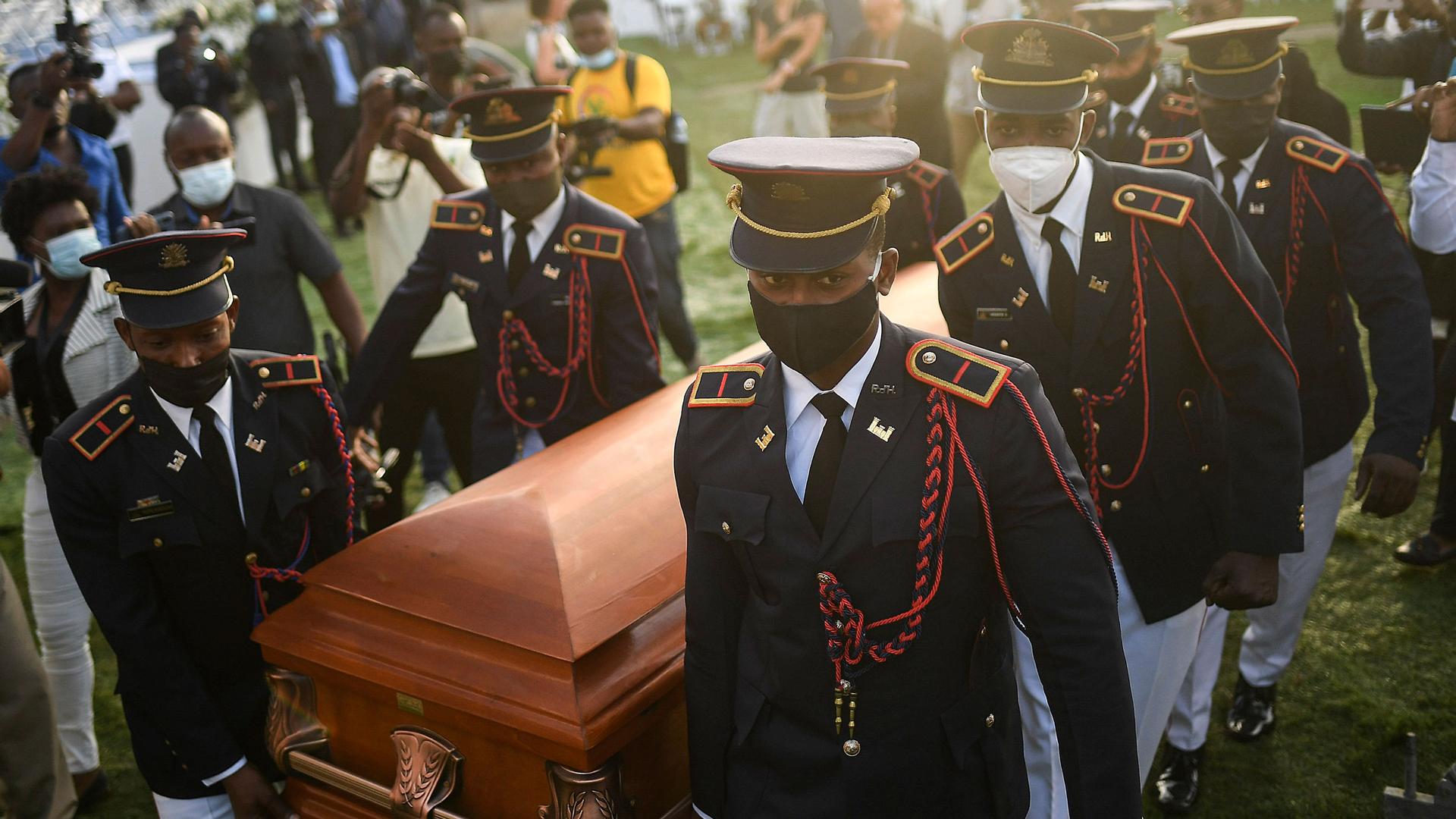 Several men wearing dress military uniforms with service caps are shown carrying a brown wooden coffin.