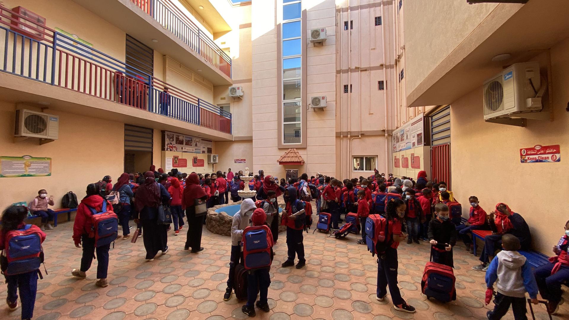 A large crowd of young students are shown wearing backpacks and walking in a courtyard with tan-colored buildings on three sides.