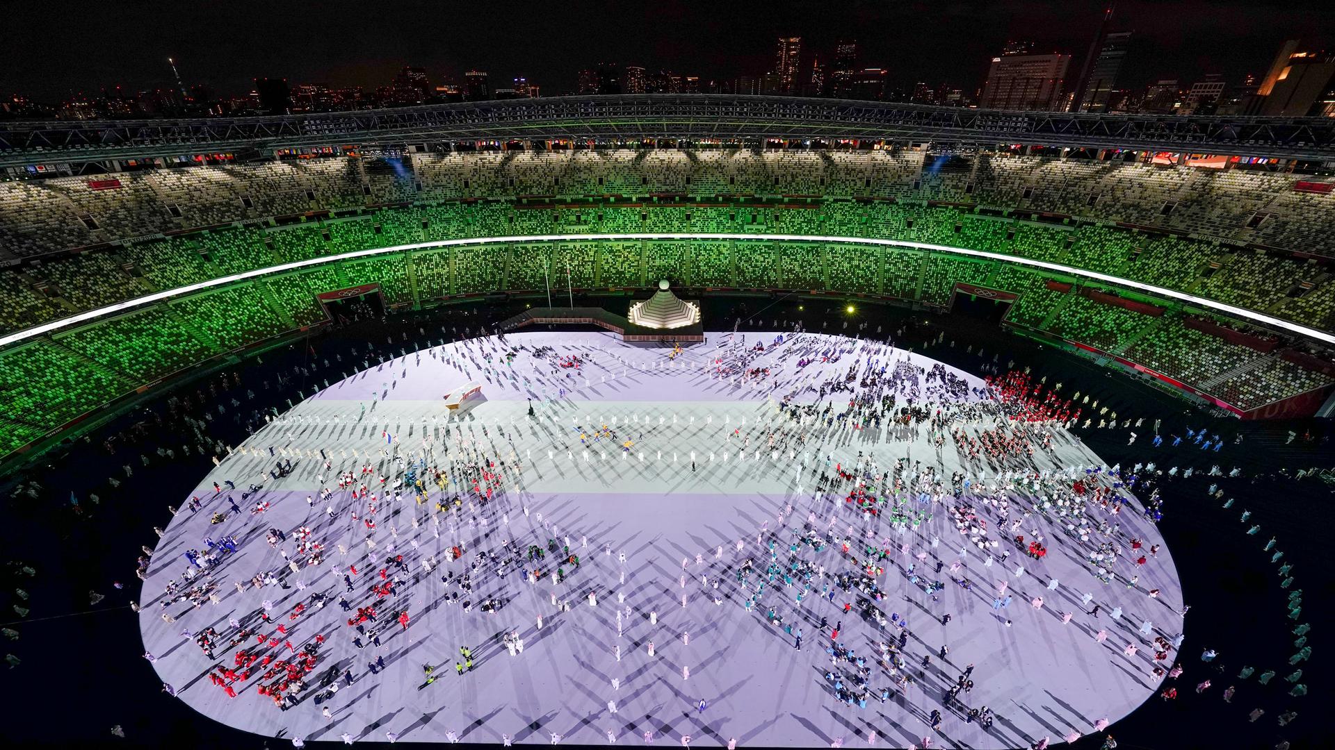 Olympic athletes are shown on the white floor of a large oval stadium shown at night with colorful lighting and mostly empty seats.