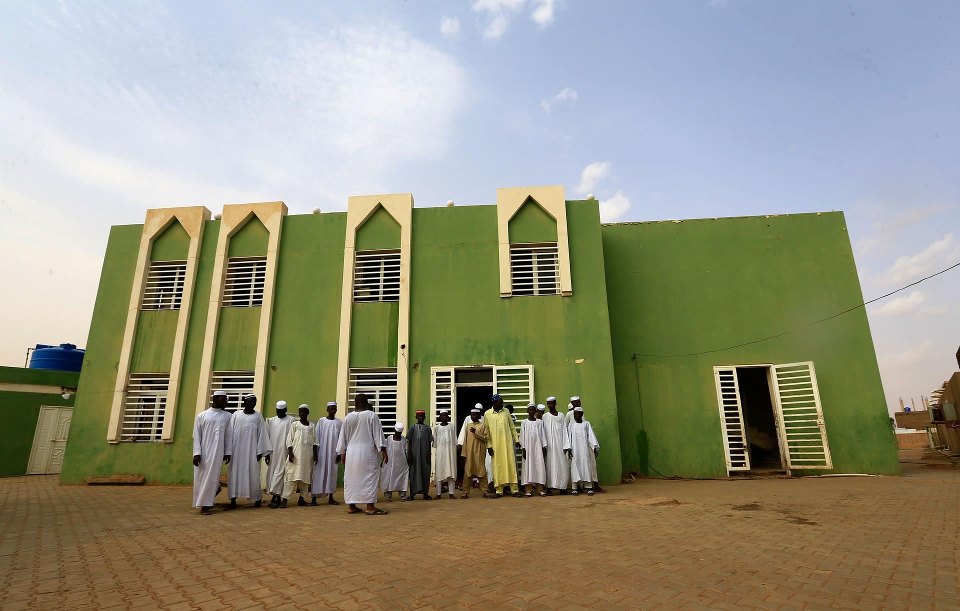 A group of men are shown in traditional Islamic robes outside of the green facade of the Haj Yousif Mosque.
