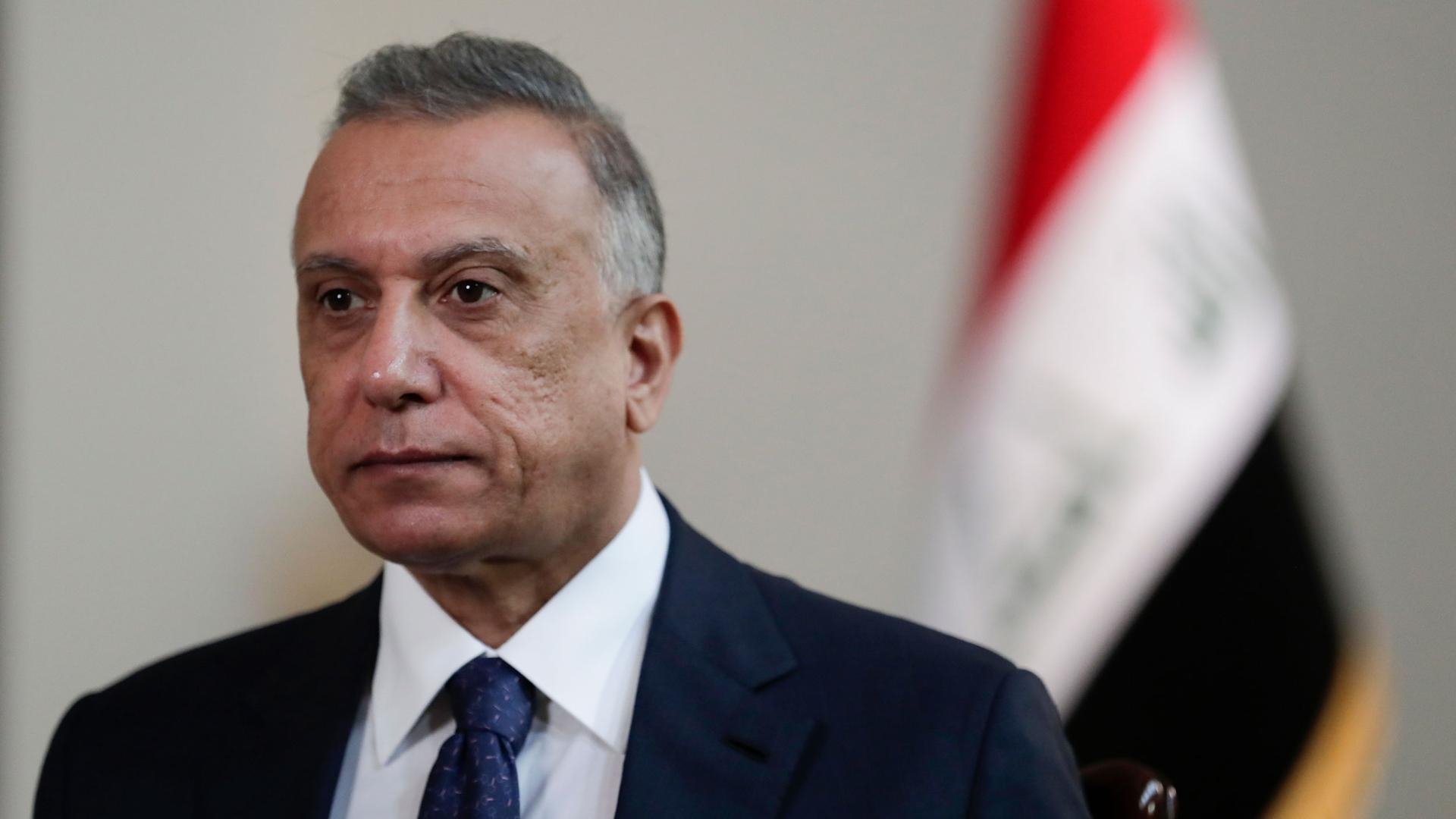 Iraqi Prime Minister Mustafa al-Kadhimi is shown wearing a dark suit and tie with the Iraqi national flag in soft focus in the background.