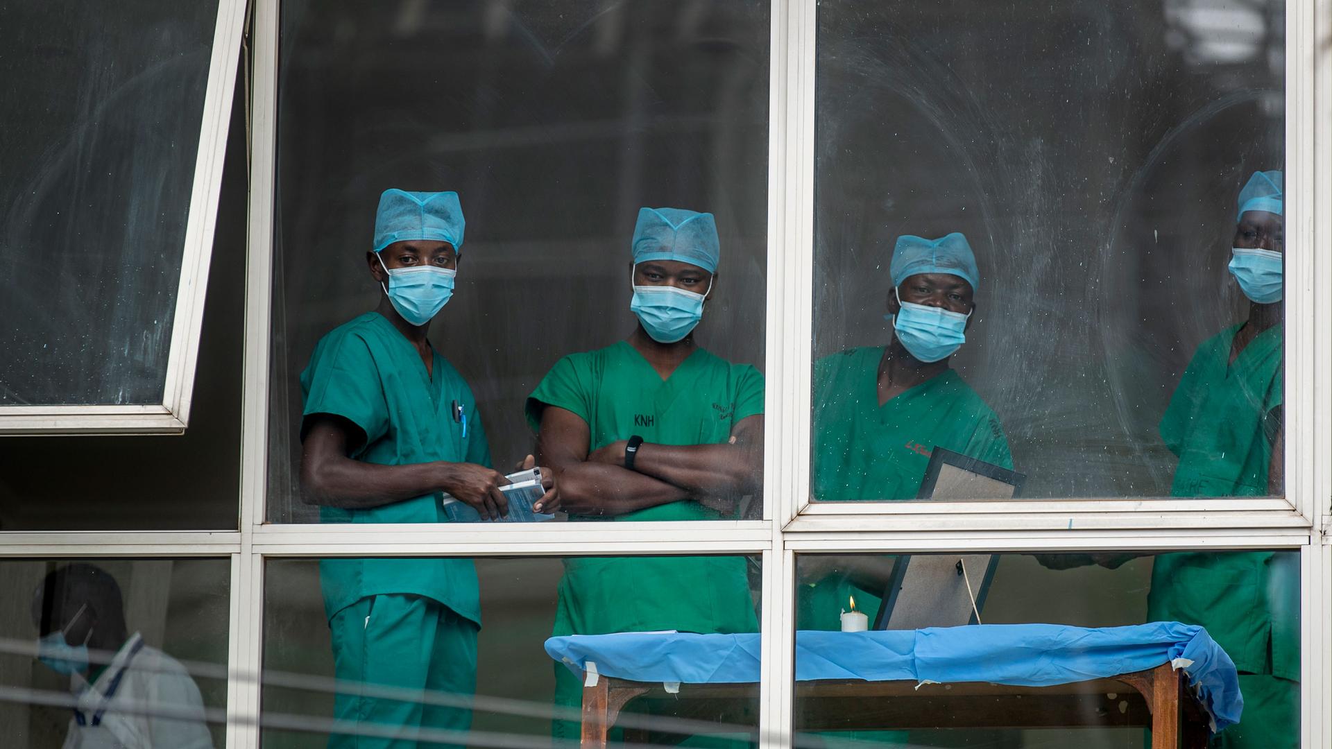 Four people are shown looking through a white-framed windows and wearing green medical scrubs.