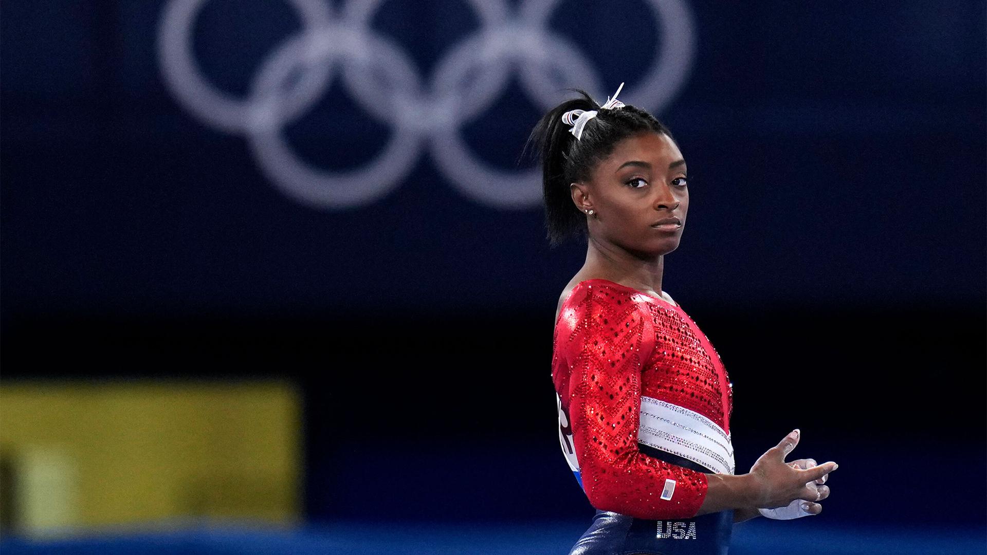 Simone Biles is shown in a profile photograph looking to her right, wearing a red, white and blue uniform with the Olympic rings in the distance.