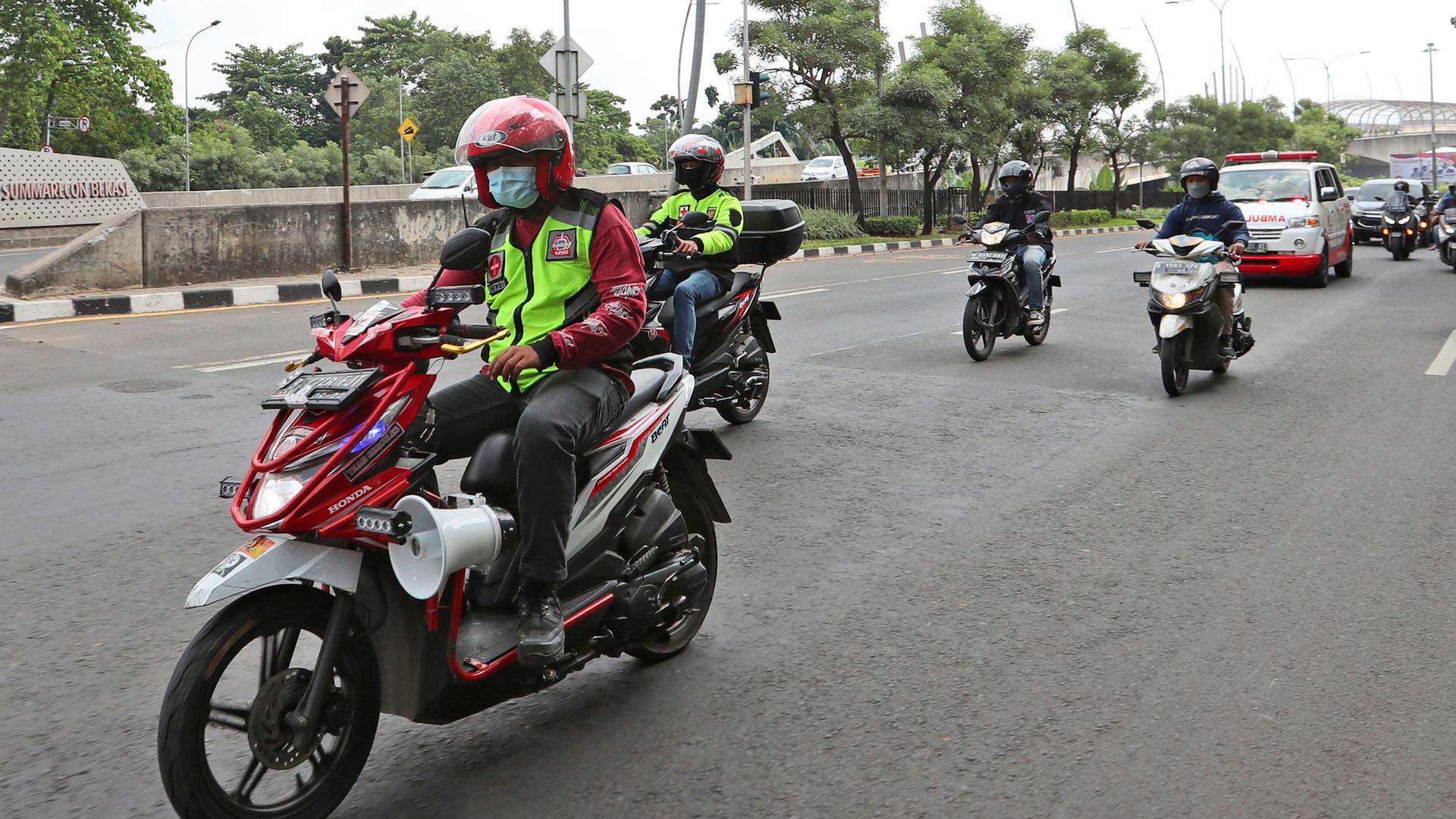 Several people on motorbikes and wearing helmets are shown riding in front of an ambulance. 