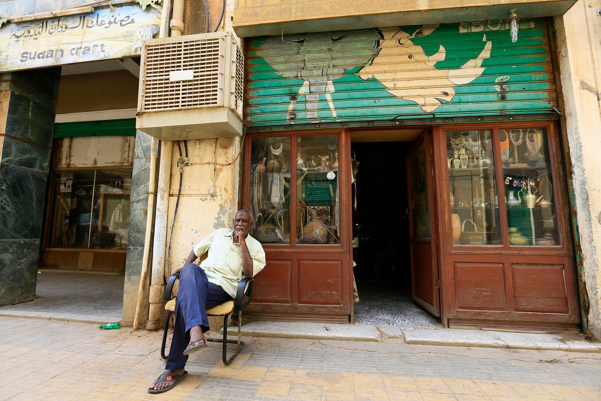 A man is shown sitting with his legs crossed in a chair outside of a shop with windows diplaying antiques.