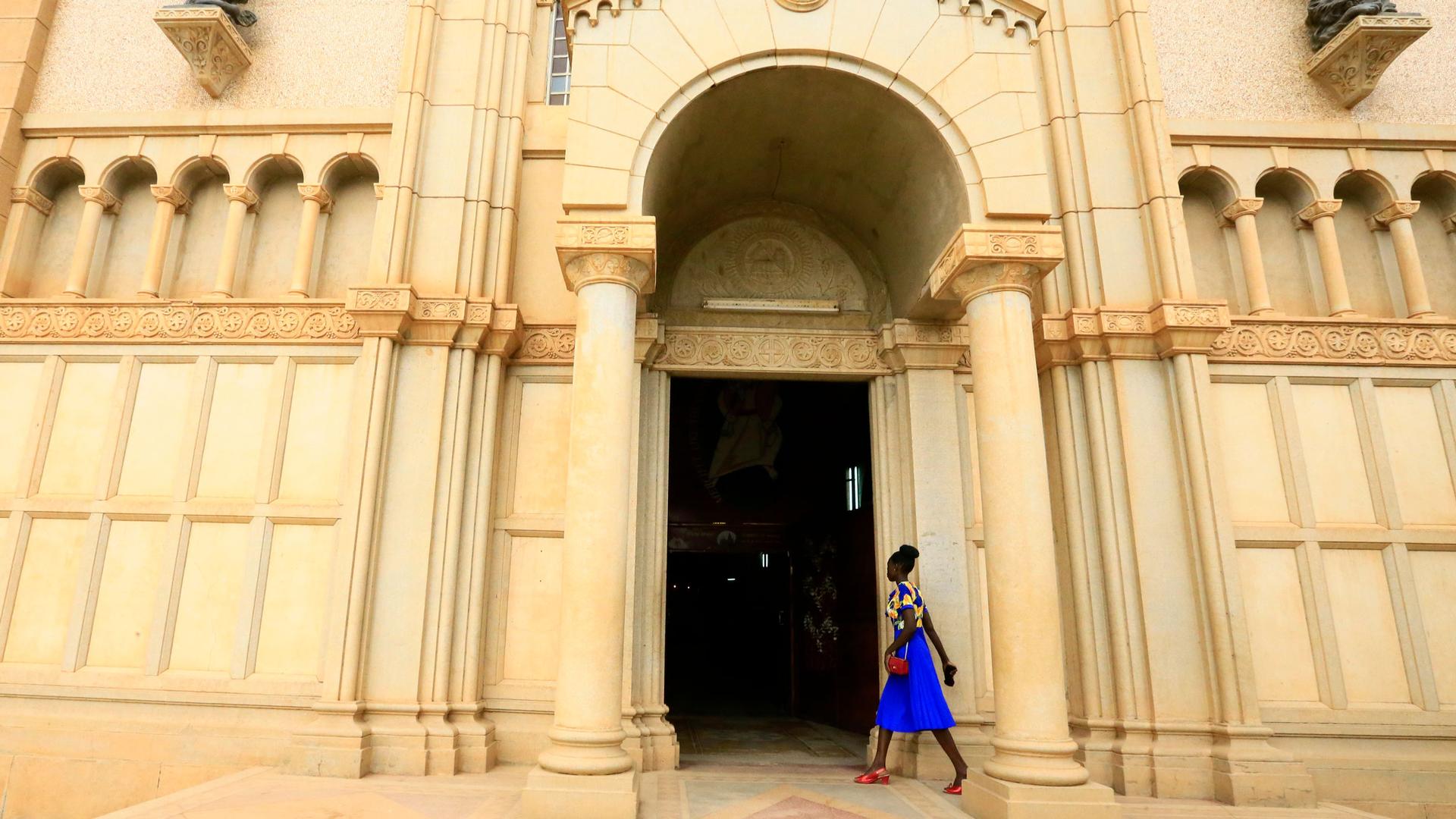 A woman wearing a blue dress and red shoes walks into the tan-colored stone entrance to St. Matthew’s Catholic Cathedral