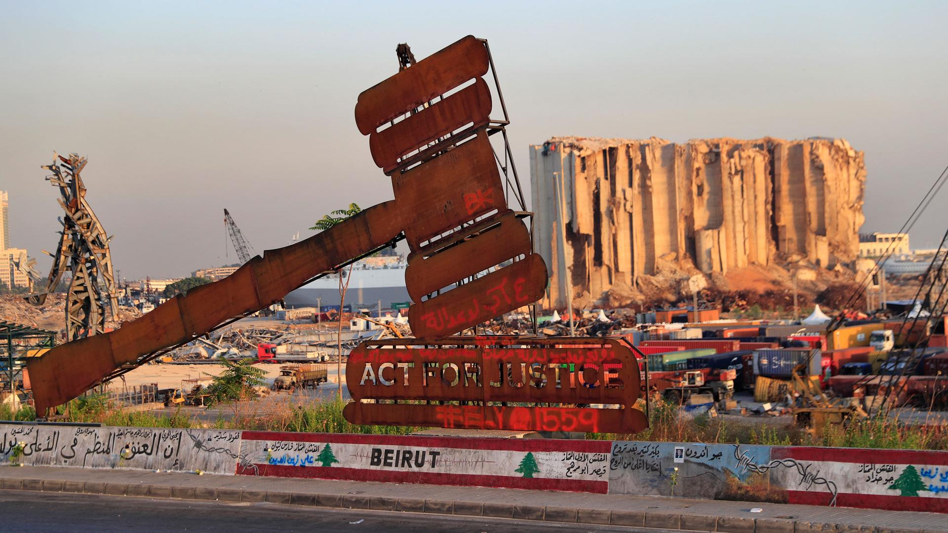 A large gavel made of metal is shown in the near ground hammering a sign that reads "Act for Justice" with a bombed out grain silo in the distance.