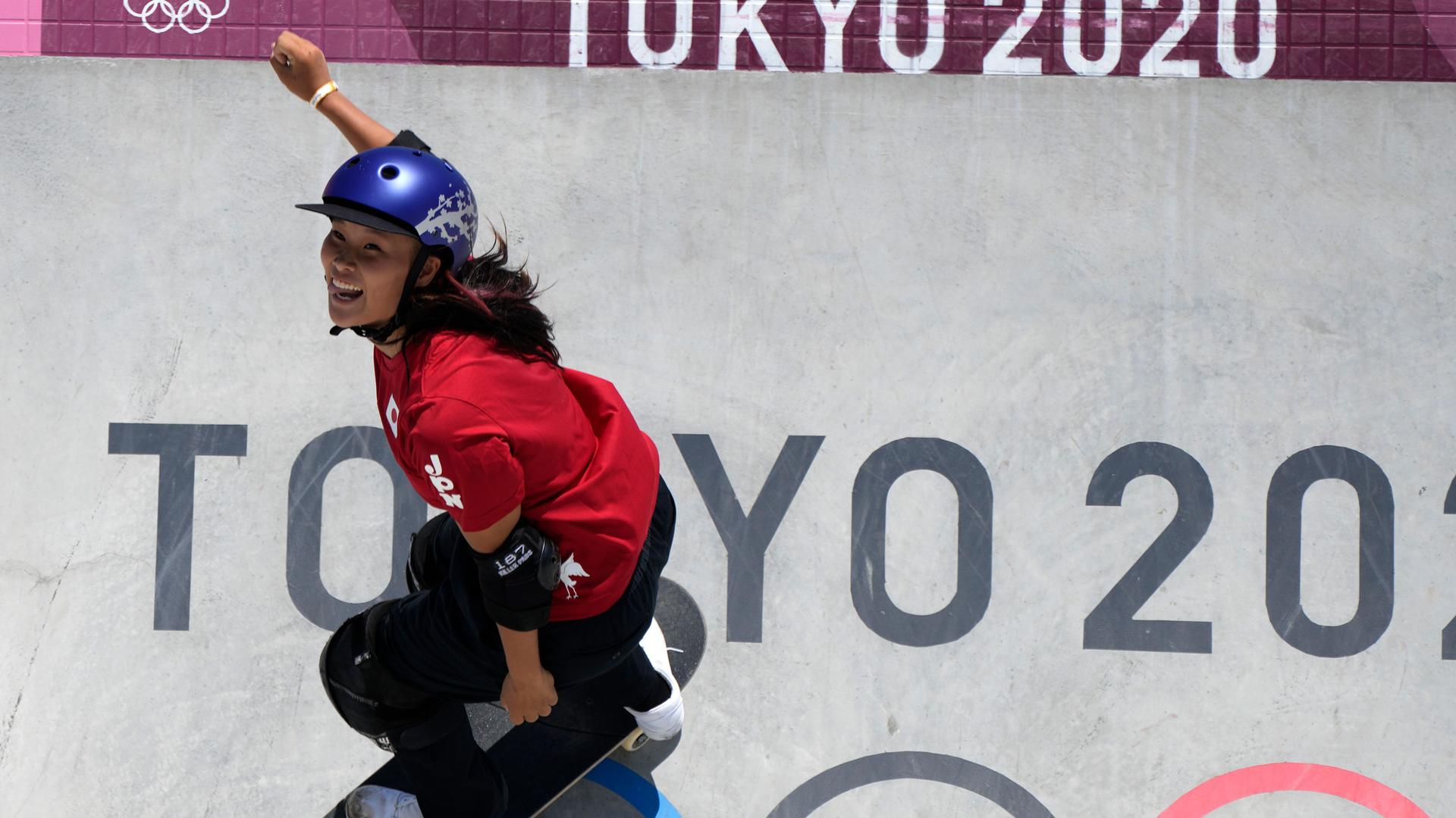 Sakura Yosozumi is shown wearing a red shirt and purple helmet while skateboarding down a wall with "Tokyo 2020" painted on it.