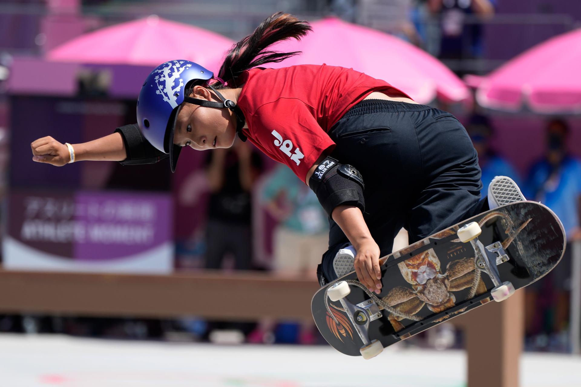 Sakura Yosozumi is shown in mid-air holding her skateboard to her feet.