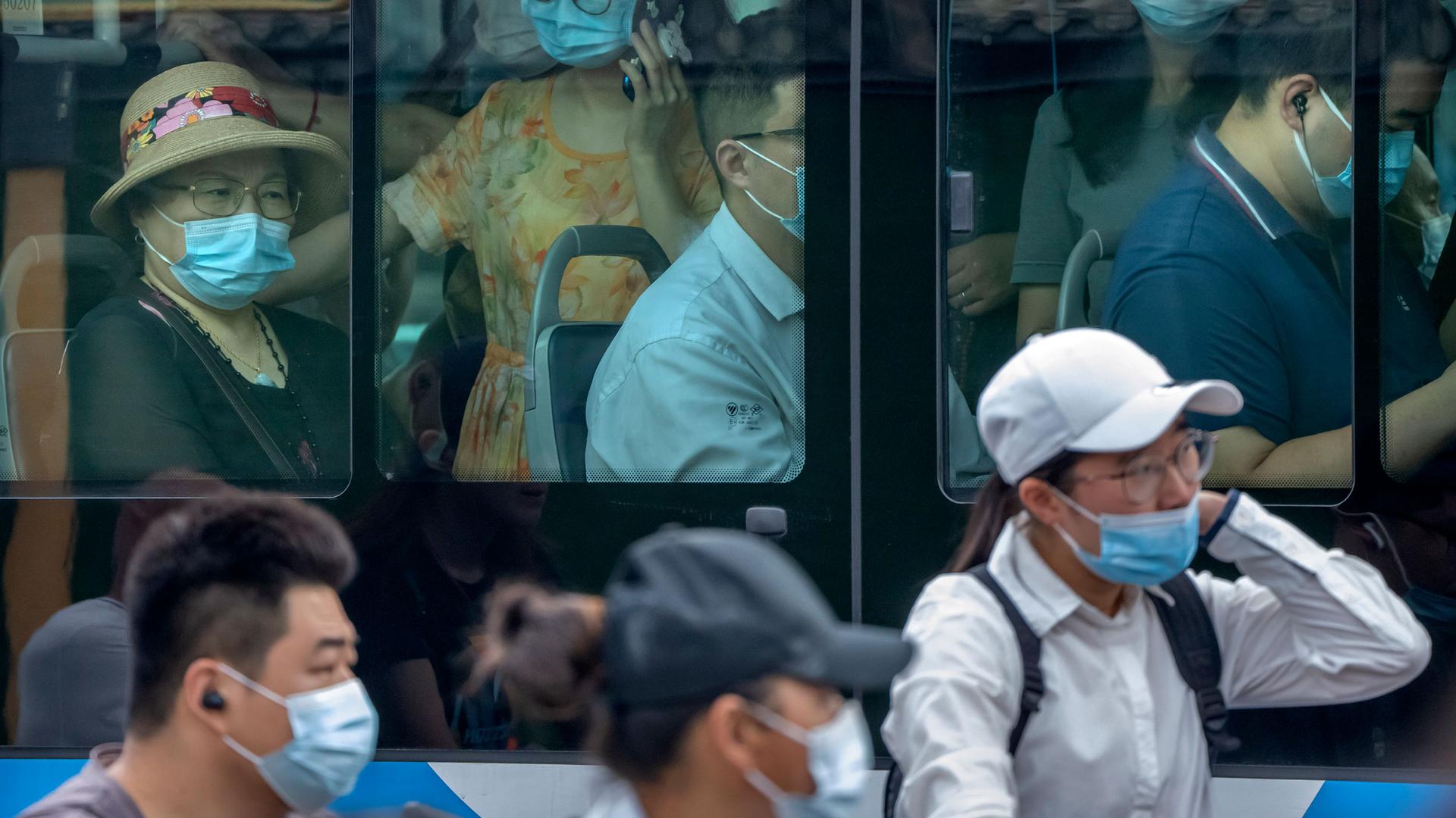 Several people are shown through the window's of a crowded bus with many wearing blue medical masks.