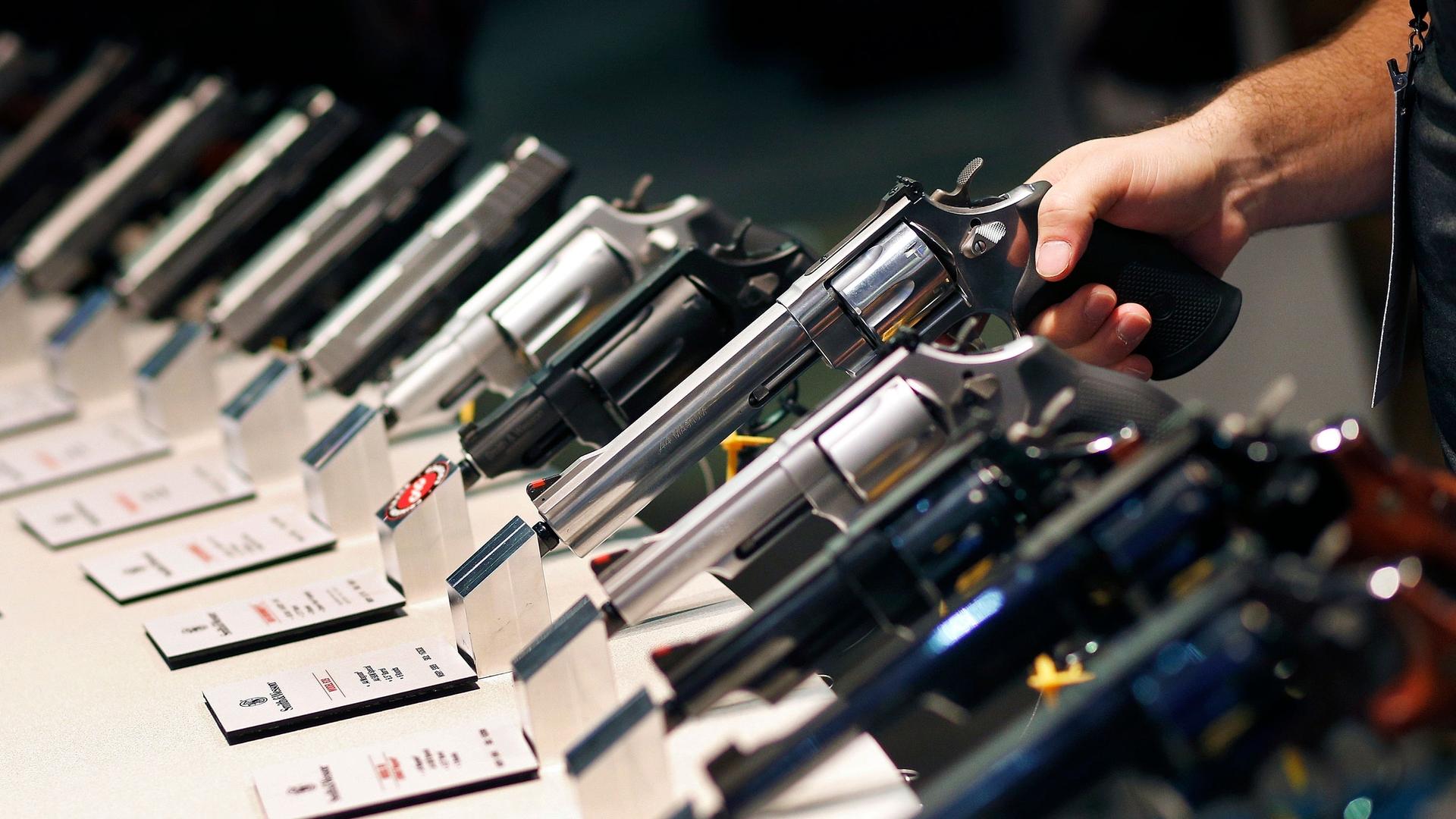 A row of handguns are shown with the handle facing outward and a hand holding on to one of the weapons.