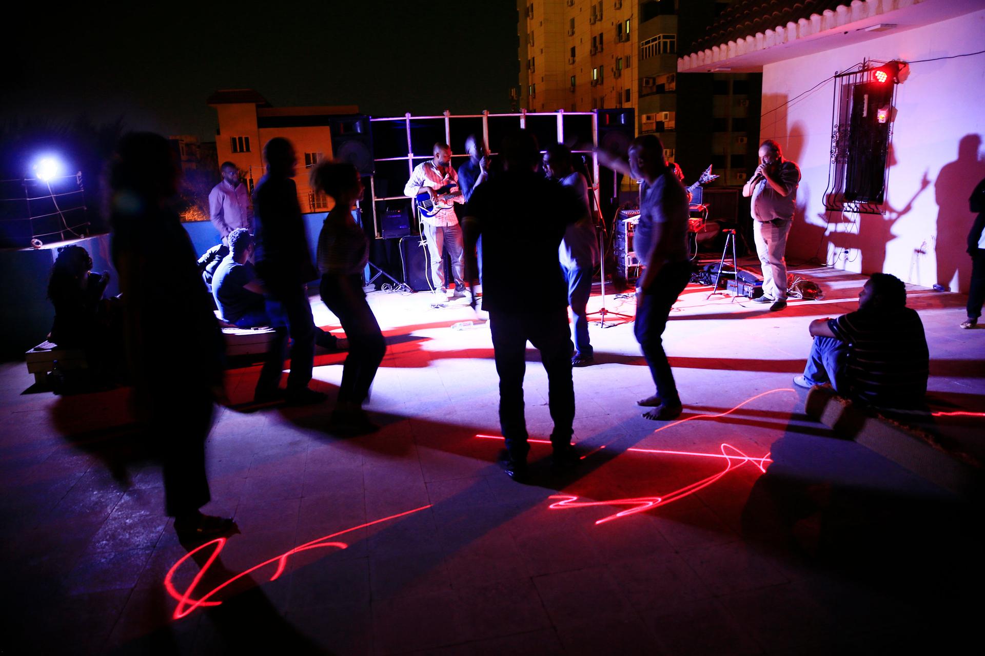 A crowd of people are shown in purple and red lighting, dancing with a band playing in the distance.