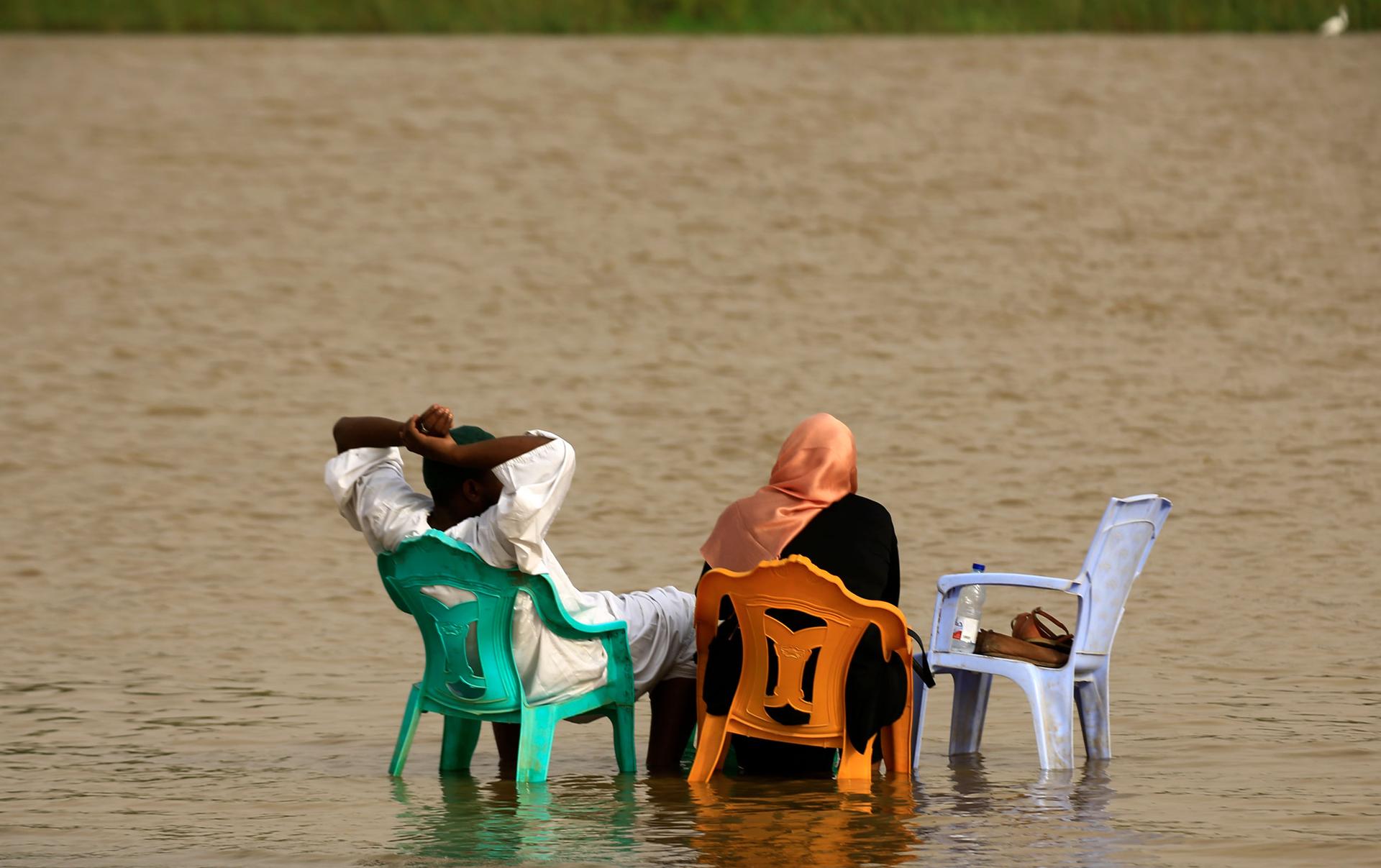 A man and woman are shown from behind both sitting in plastic chairs submerged in the water to their ankles.