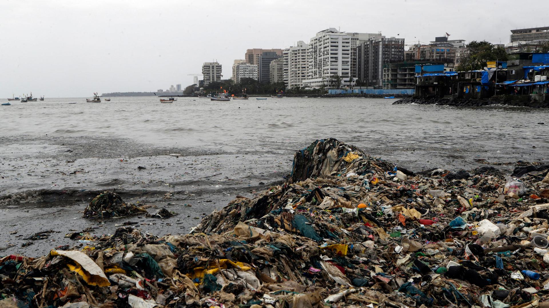 A large pile of garbage is shown along the shores of Mumbai, India, with several tall buildings in the distance.