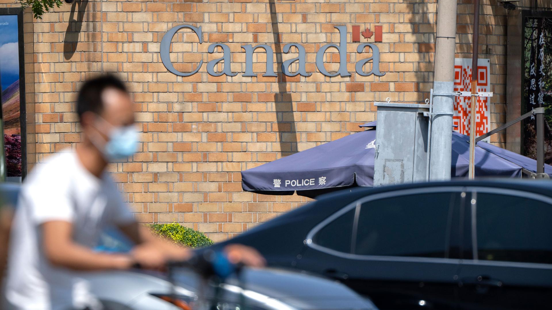 A man is shown riding a scooter in blurred focus while wearing a facemask with the brick facade of the Canadian embassy building in the distance.