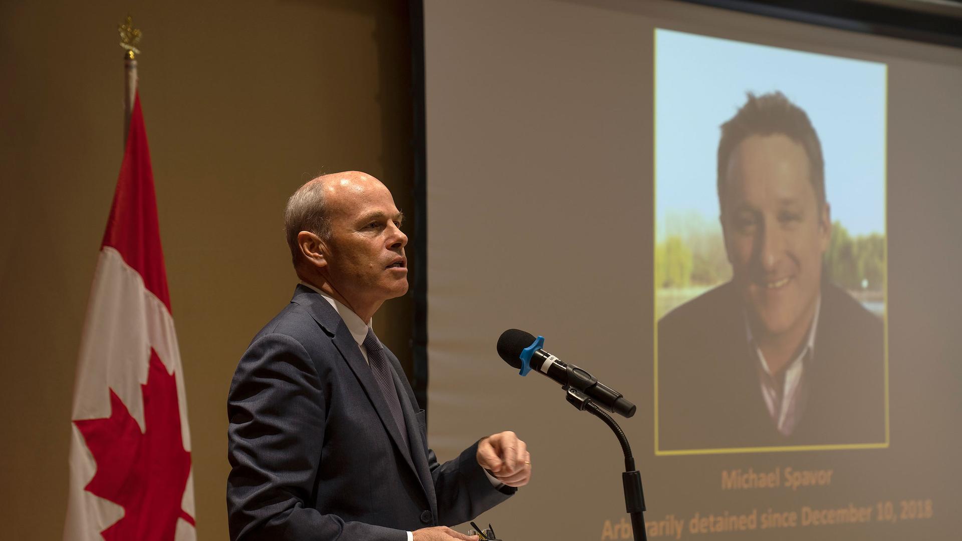 Jim Nickel is shown wearing a suit and tie while speaking into a microphone with an image of Michael Spavor projected on a screen in the background.